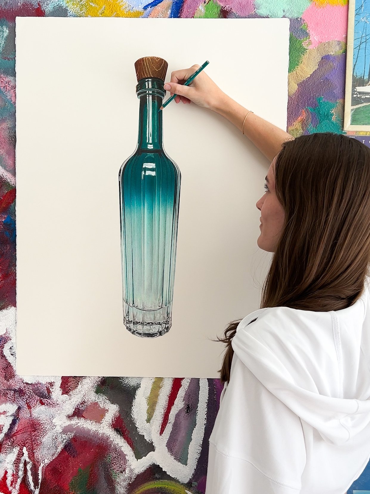 A woman painting a large, detailed image of a vintage glass bottle with a cork on a white canvas in an artist's studio.