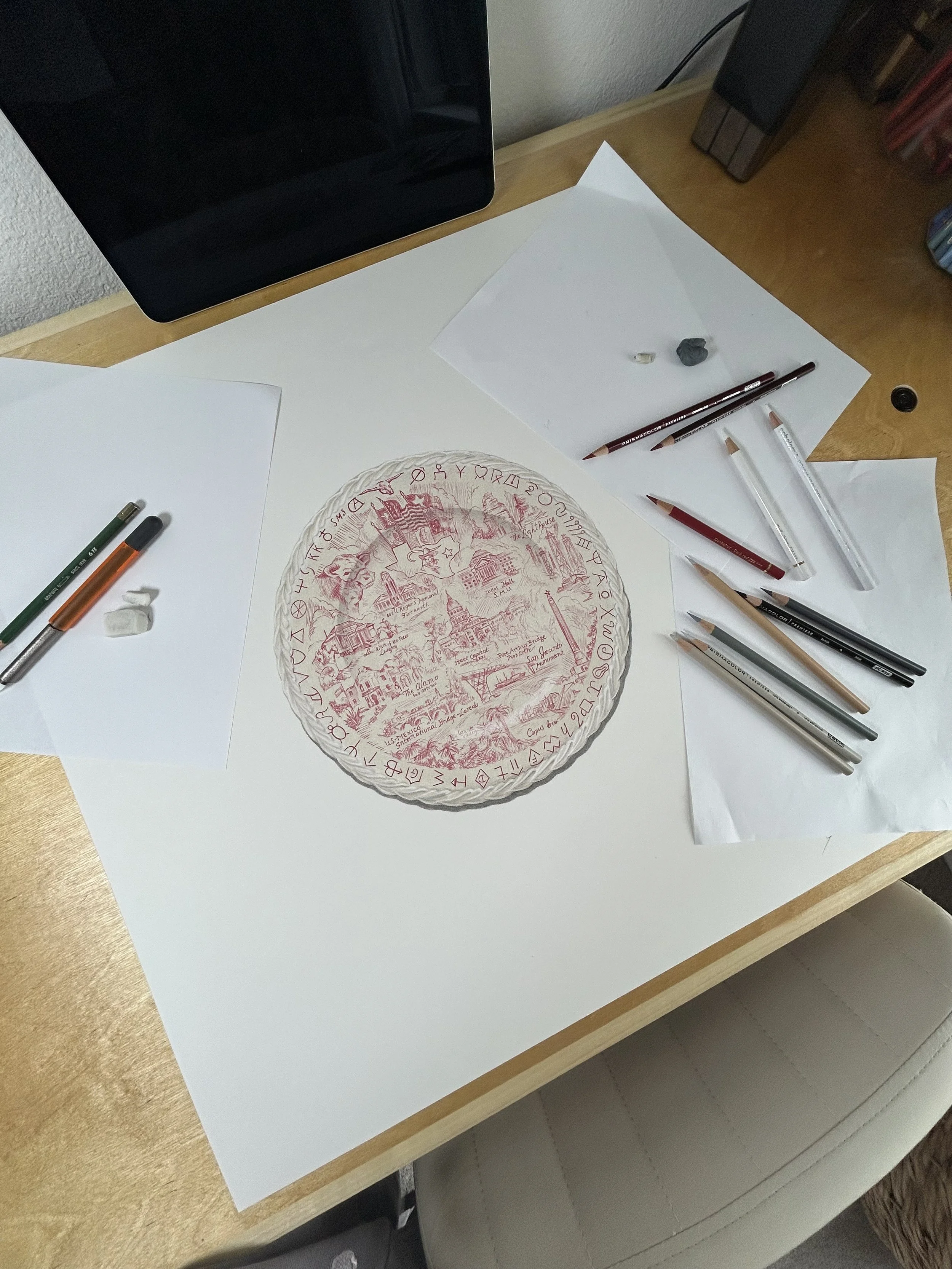 A white sheet of paper on a wooden desk with a detailed red ink drawing of a circular scene, surrounded by mysterious symbols. Various art supplies, including pens, pencils, and erasers, are nearby on the desk.