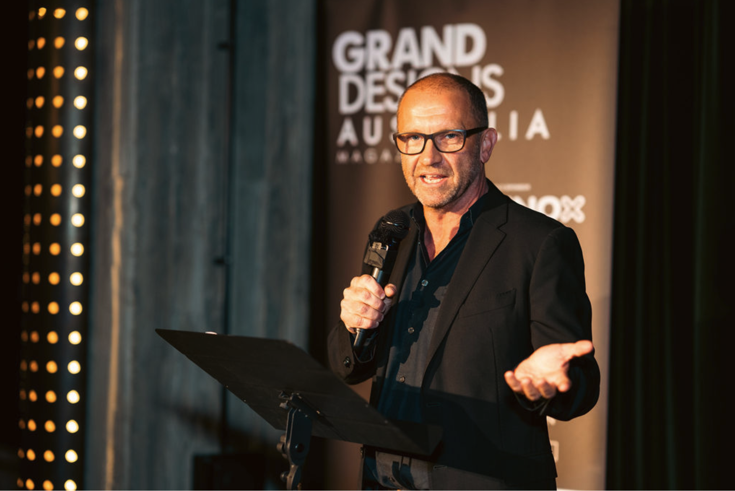Anthony Burke wearing a black suit, holding a microphone on stage with a Grand Designs Australia backdrop