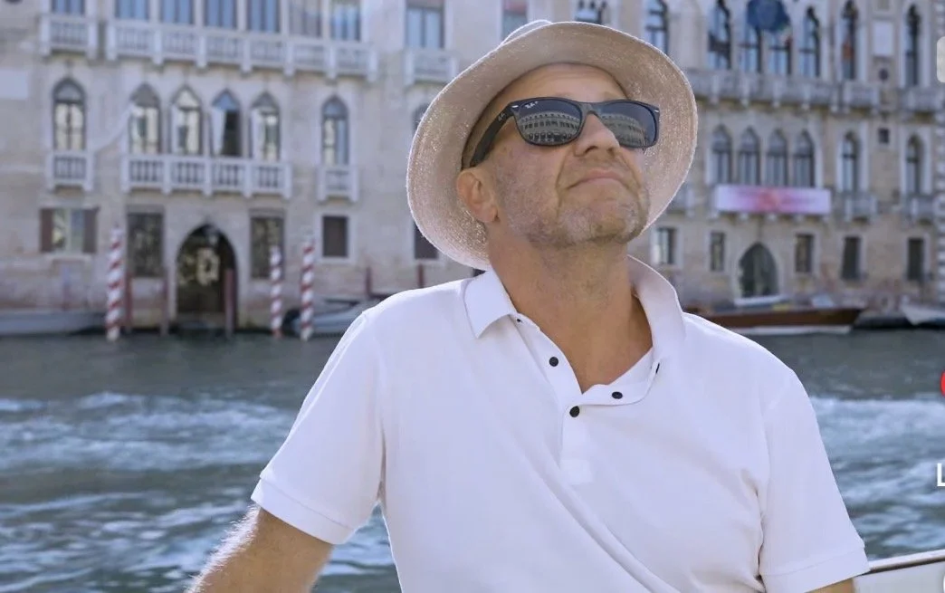 Anthony Burke on a boat in the Venice Lagoon wearing a hat and sunglasses looking up at the buildings