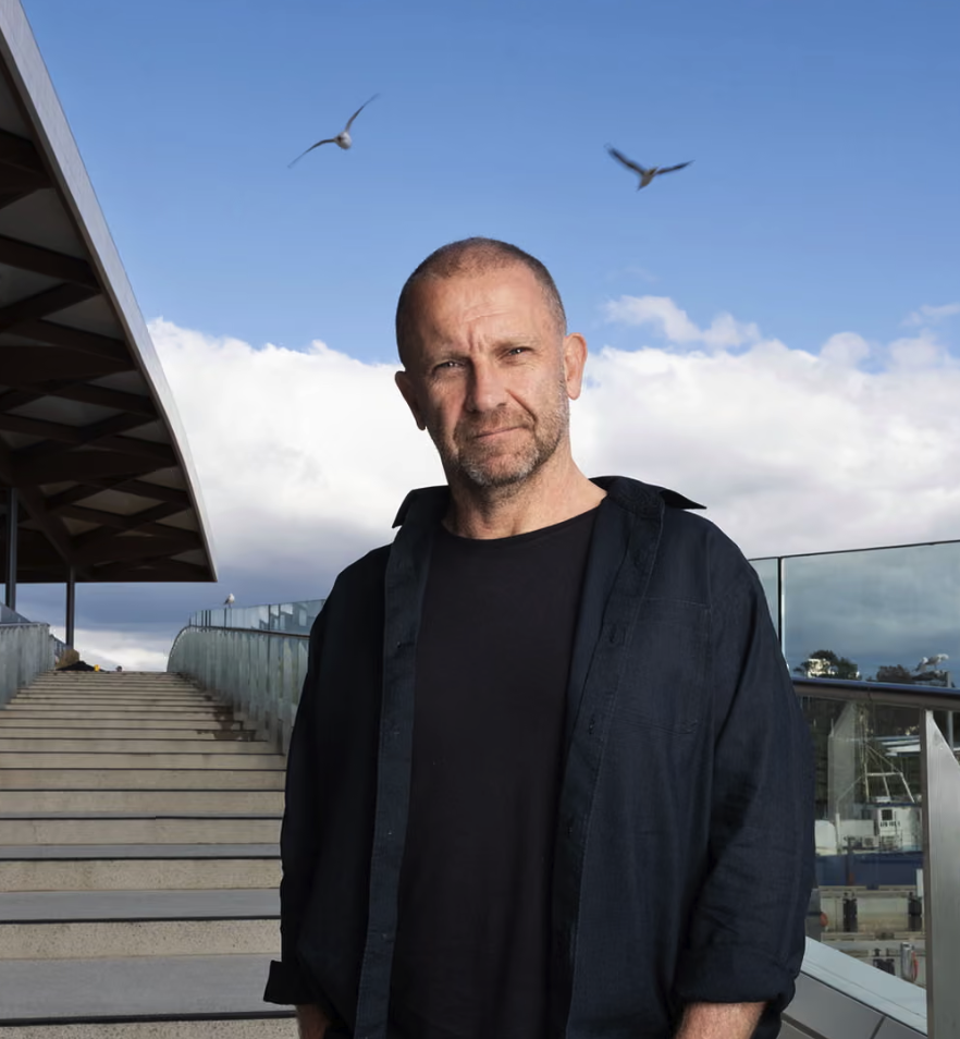 Anthony Burke standing with his hands in his pockets, wearing a black t-shirt and open shirt at the steps of the Sydney Fish Markets. Two seagulls are flying in the blue sky above.