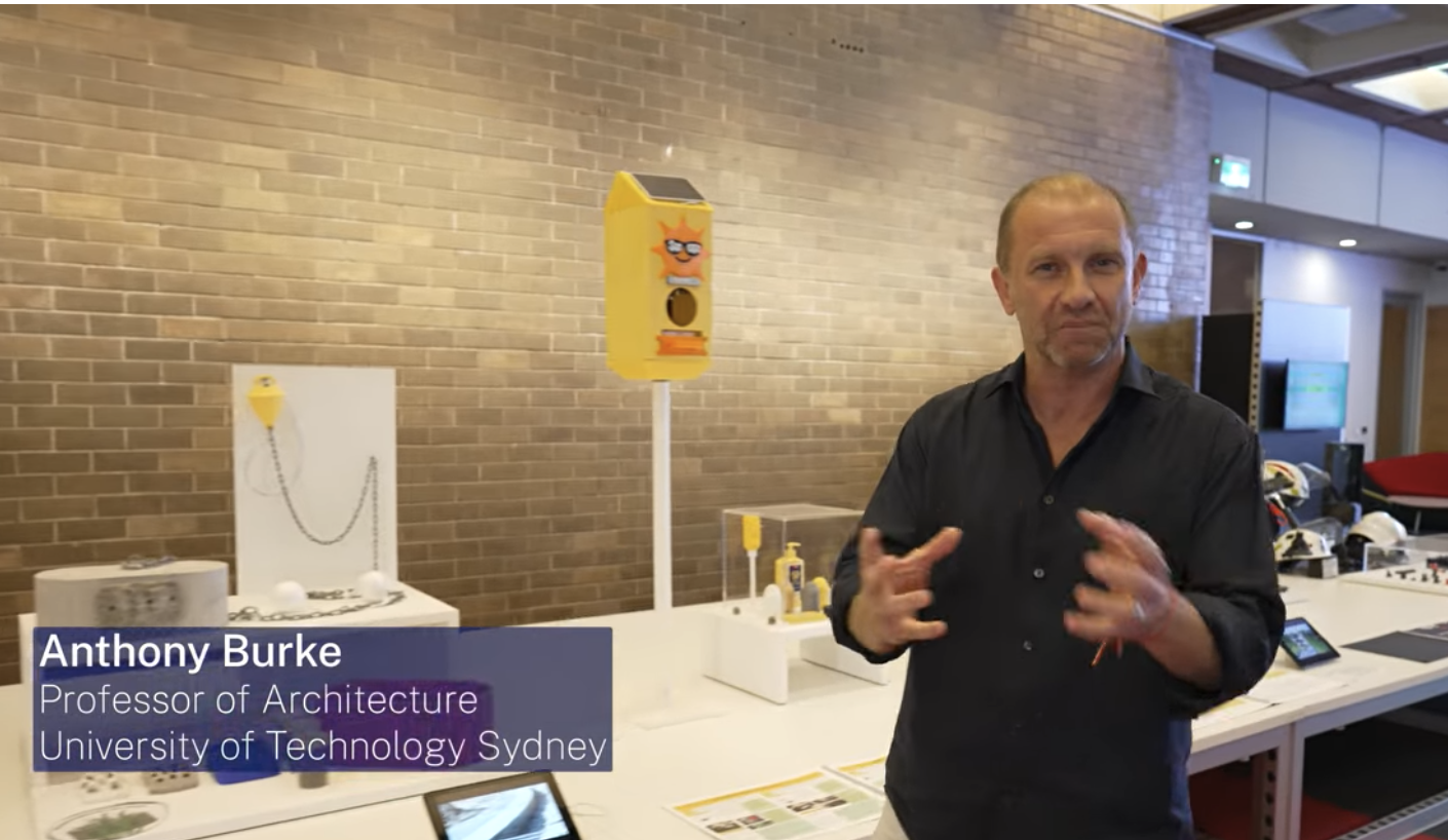Anthony Burke wearing a dark shirt in front of table which shows product prototypes, looking at the camera