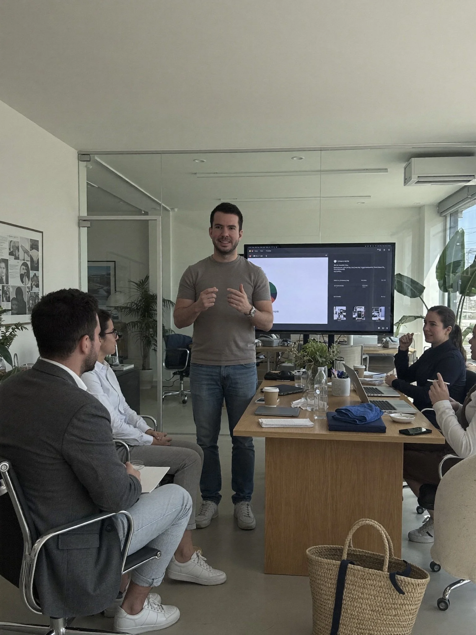 A man in a gray T-shirt and jeans is speaking to a group of diverse people in a modern office conference room, with a large screen behind him displaying a pie chart and social media content. The attendees are seated around a wooden table with laptops, notebooks, and drinks. A basket is visible in the foreground, and the room has large windows with natural light.