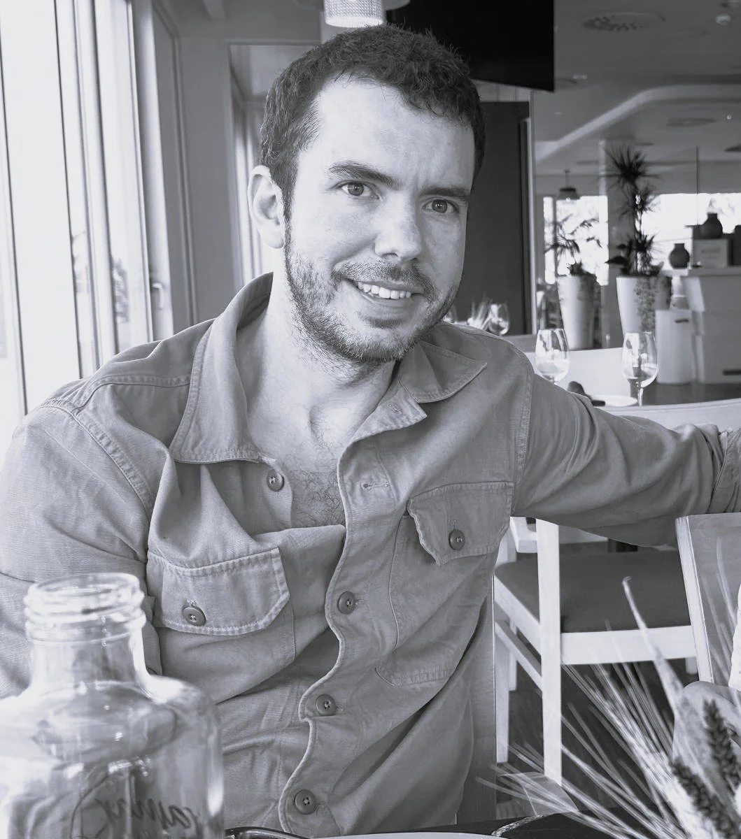 A smiling man with short dark hair and a beard, wearing a button-up shirt, sitting at a dining table in a restaurant with wine glasses and decorative plants in the background.