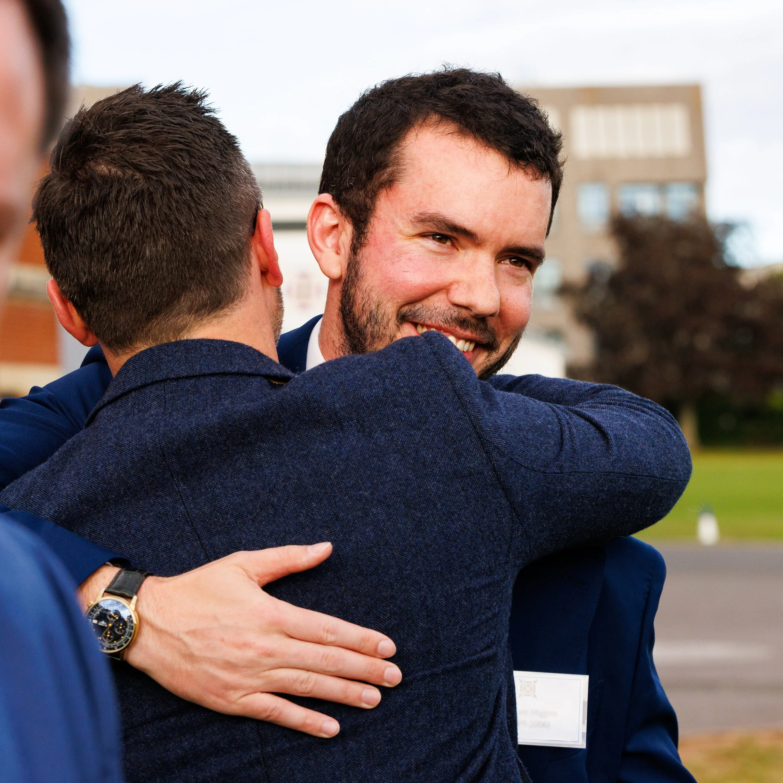 A man in a blue suit is smiling while being embraced by another person, with the person in the blue suit resting their hand on the man's chest.