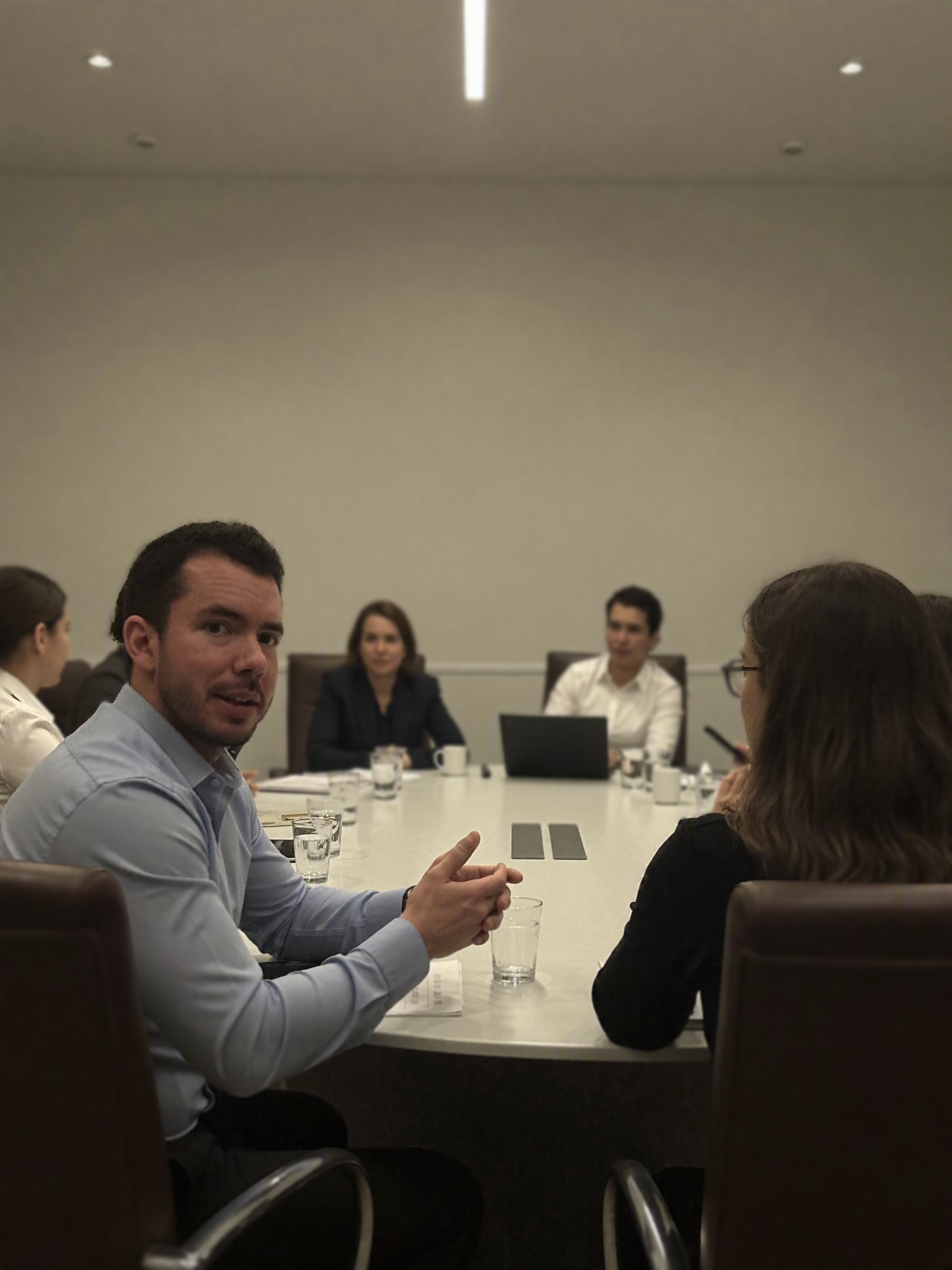 A group of business professionals sitting around a conference table during a meeting in a modern office. One man is speaking, and others are listening attentively.