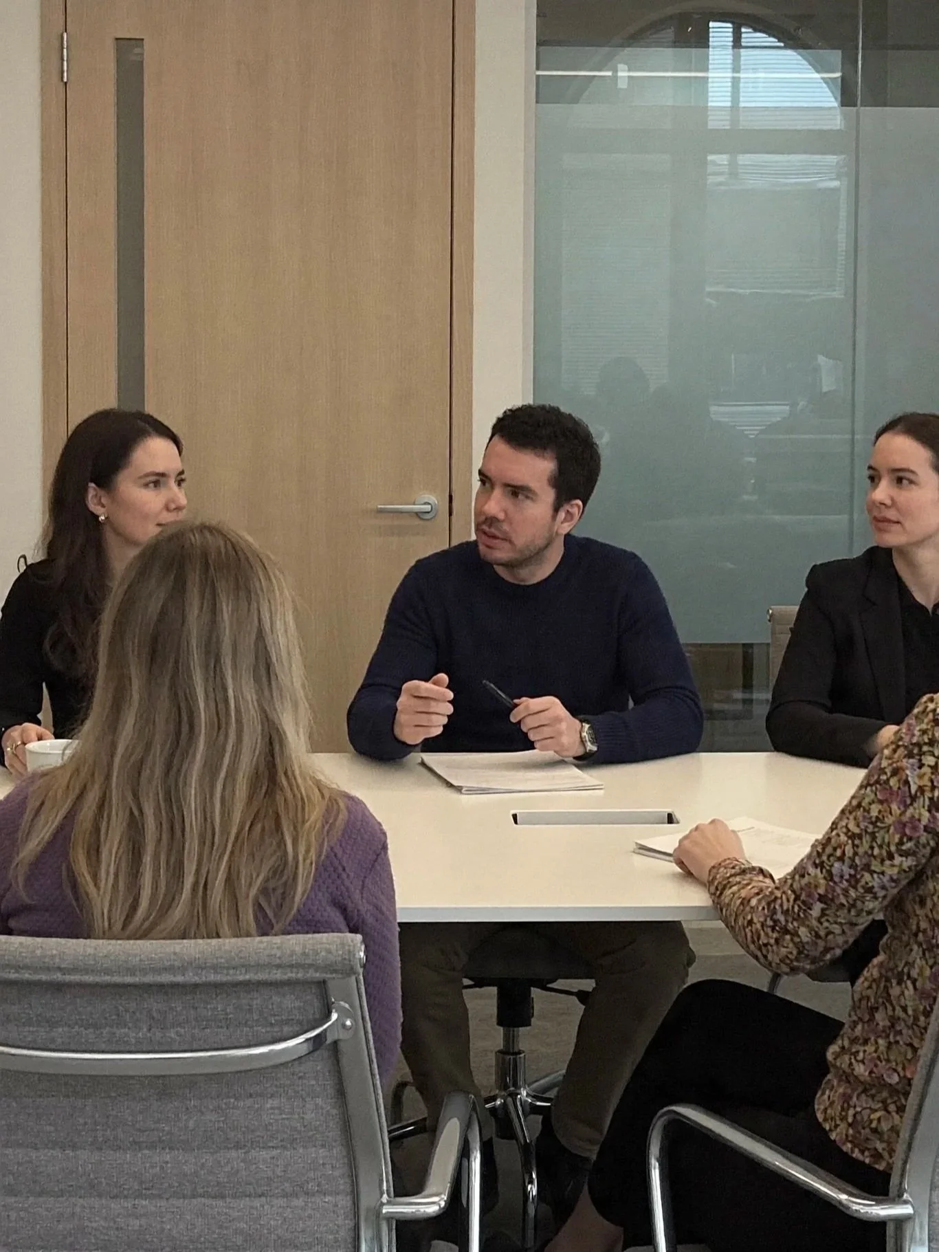 People sitting around a conference table engaged in a discussion, with a woman on the left, a man in the middle speaking holding a pen, and a woman on the right, in a modern office setting.