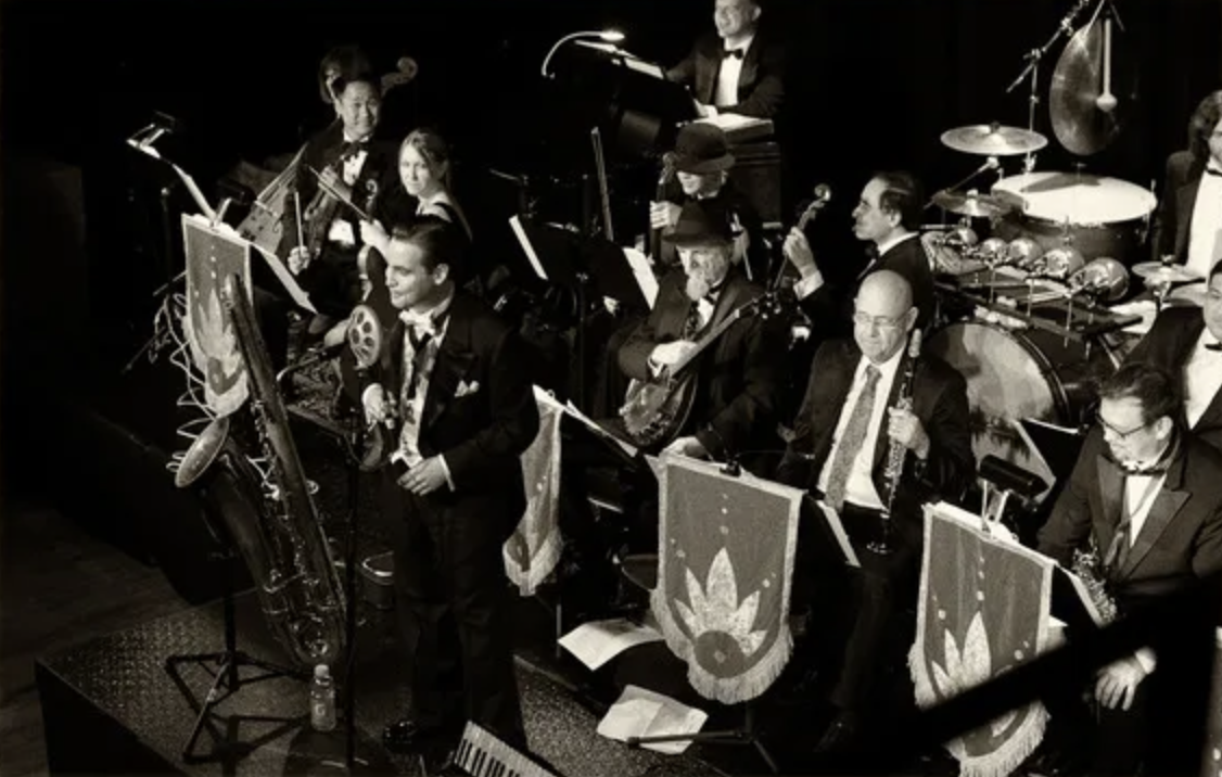 Jazz band performing on stage with musicians playing various instruments including saxophone, double bass, drums, and keyboard, some wearing tuxedos and bow ties.