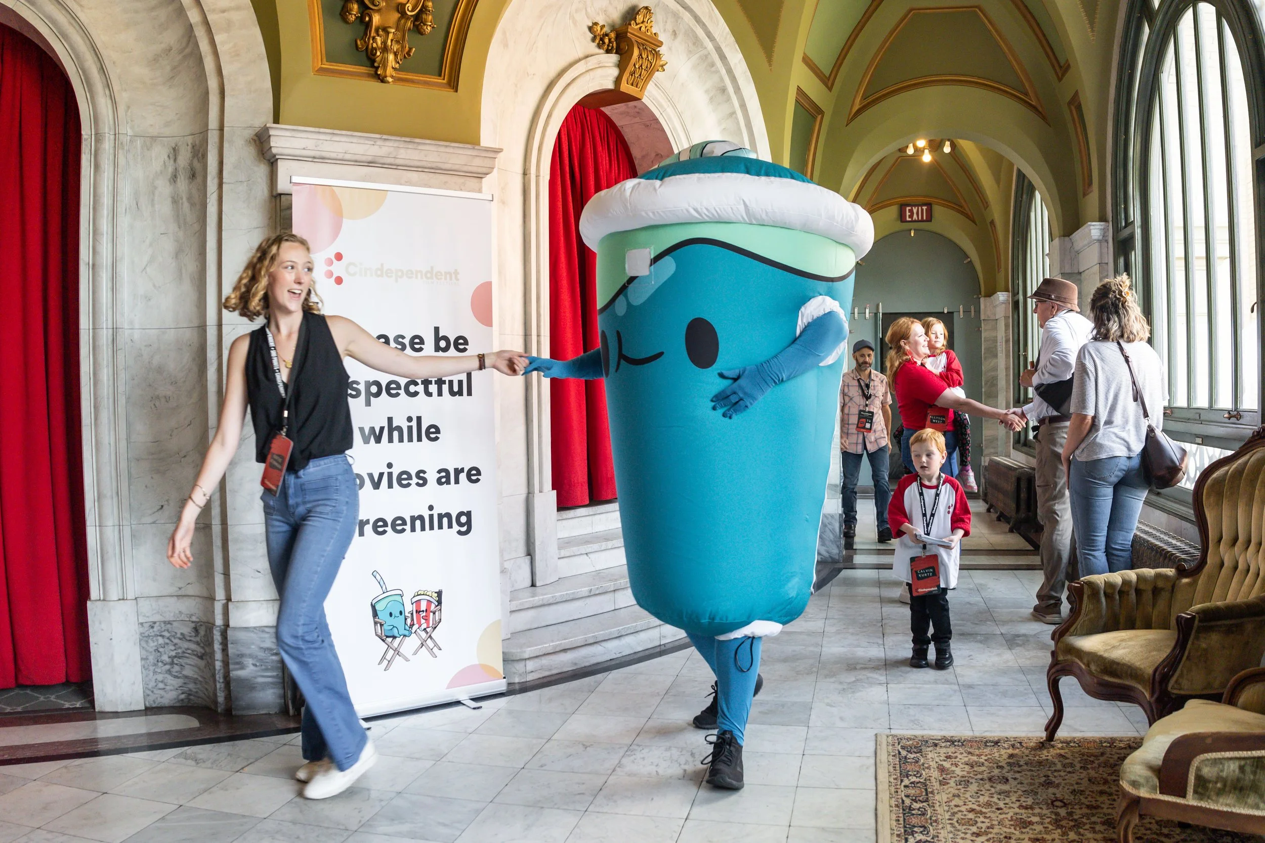Cindependent Film Festival volunteer smiling and guiding the mascot Bubs, a blue soda cup, through Memorial Hall.