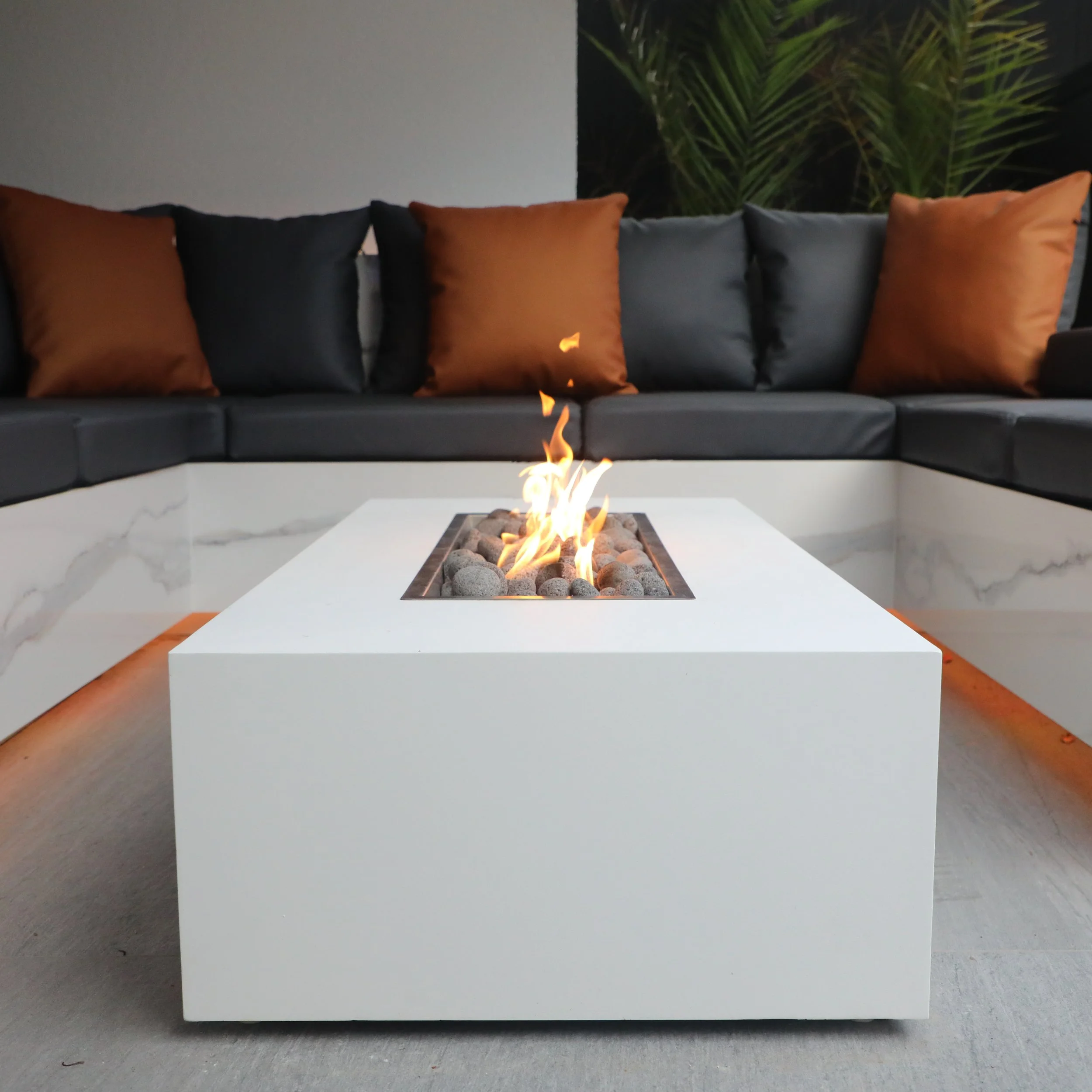 Modern outdoor seating area with a white firepit table and a black sofa with orange and black pillows, with a dark wall and greenery in the background.