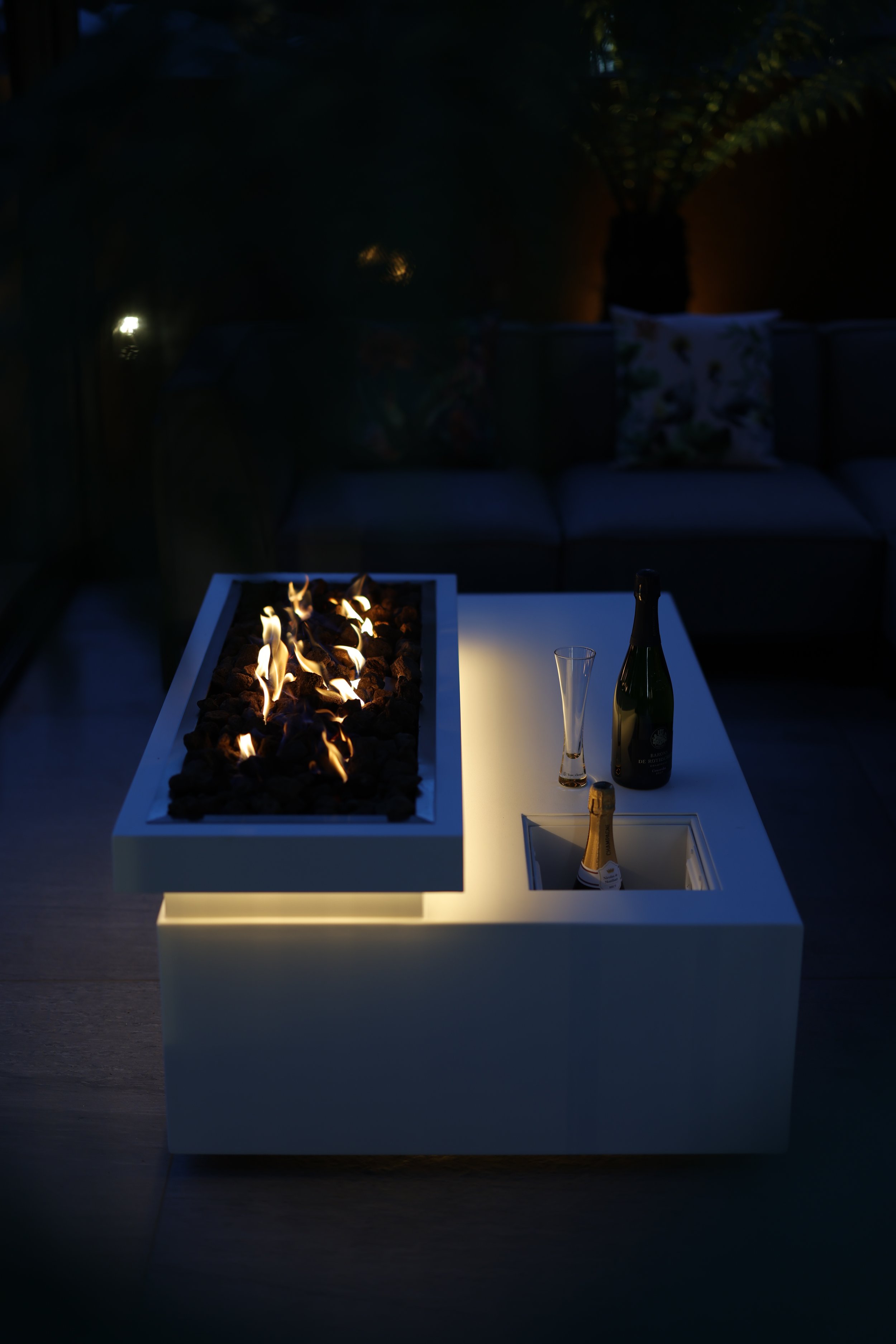 Outdoor table with a modern rectangular fire pit, a bottle of champagne, an empty champagne flute, and a glass with a drink, set at night in a backyard patio with a sofa and decorative pillows in the background.