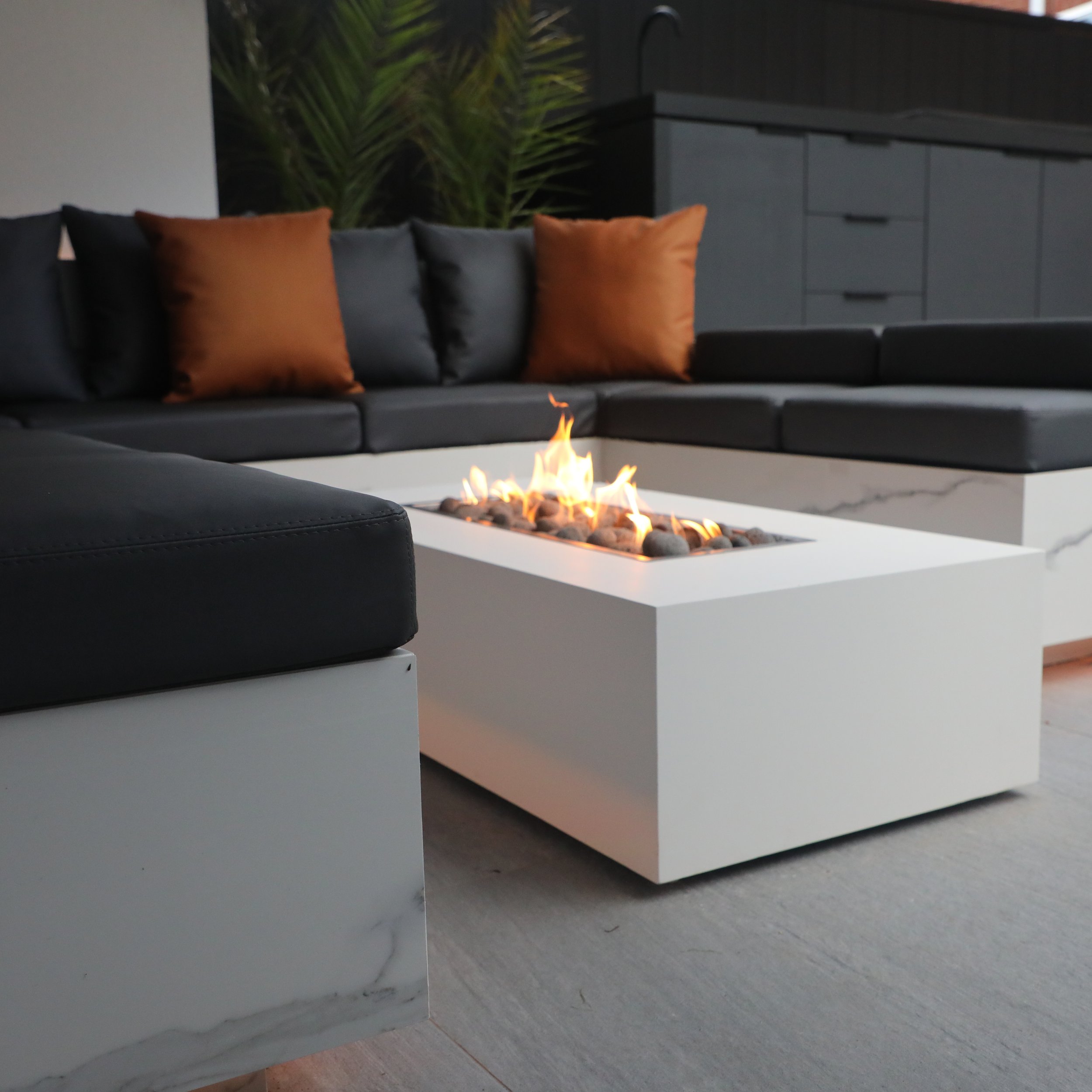 Modern outdoor seating area with black leather sectional sofa, brown pillows, and a white rectangular fire pit with flames, against a dark wall with green plant in background.