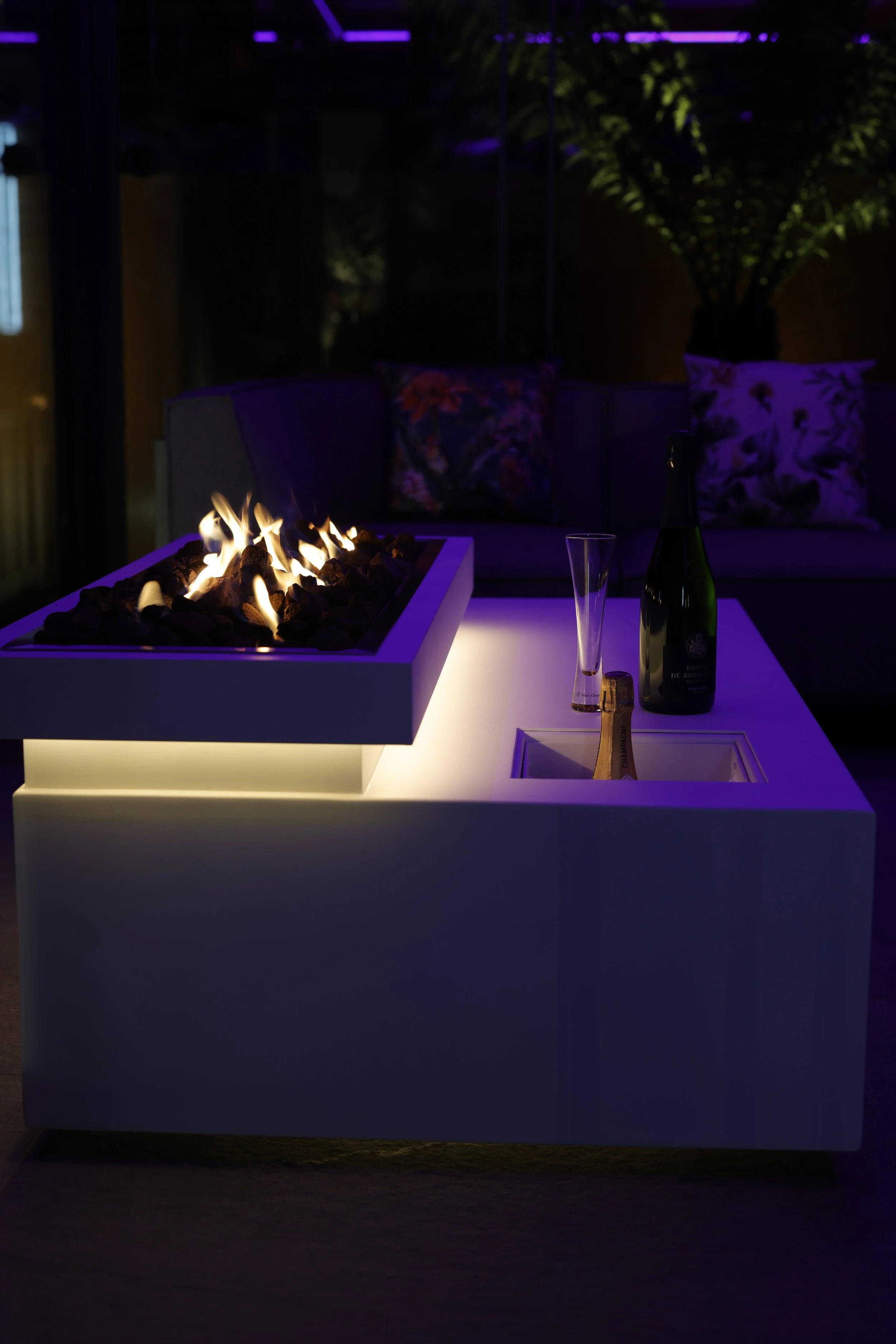 A modern white fire pit table with a built-in gas flame on top, in a cozy outdoor setting at night. There are bottles and glasses on the table, a dark sofa with patterned cushions, and green trees in the background.