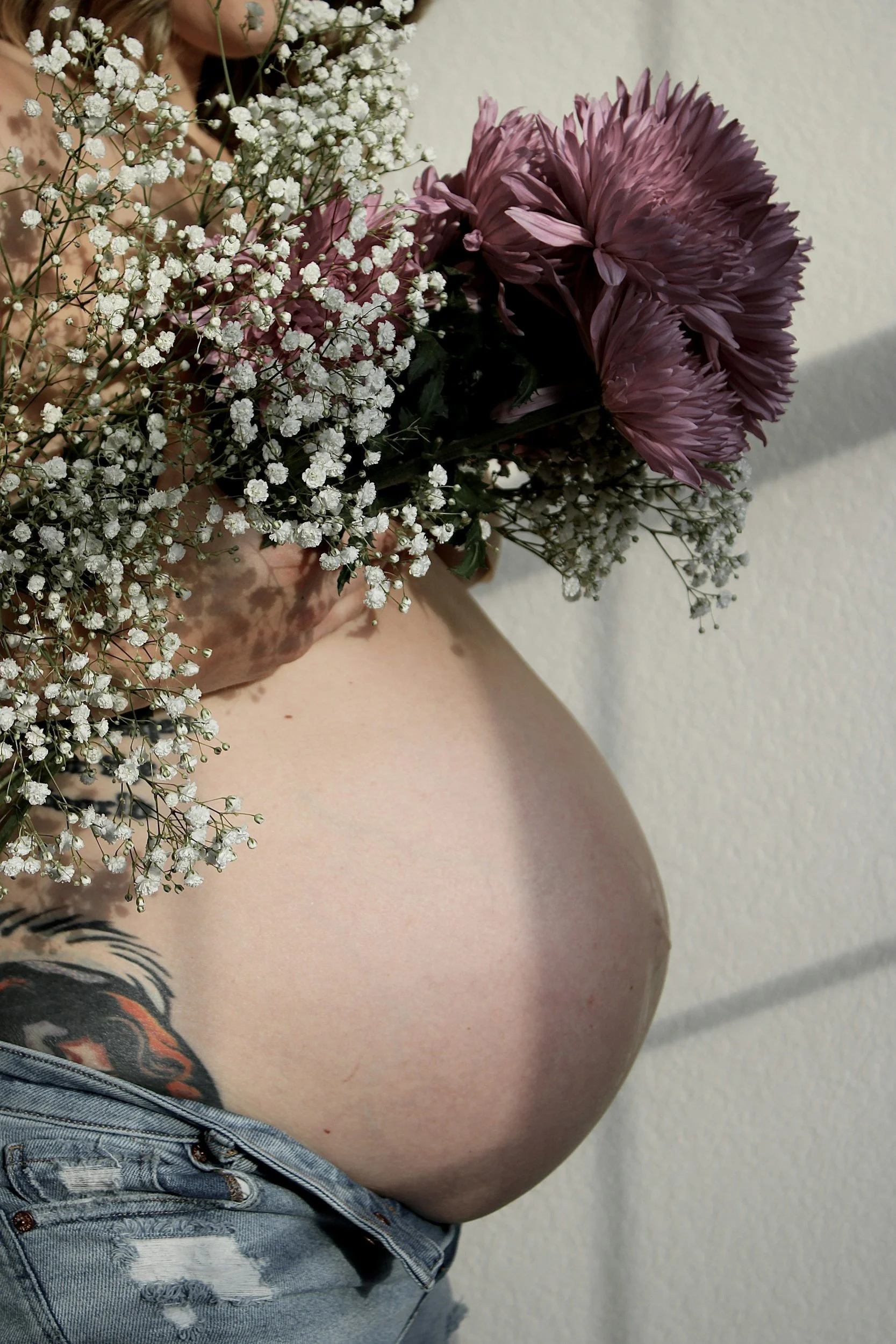 Une femme enceinte tenant un bouquet de fleurs violettes et blanches, portant un jean déchiré. La photo est prise de profil contre un mur clair.