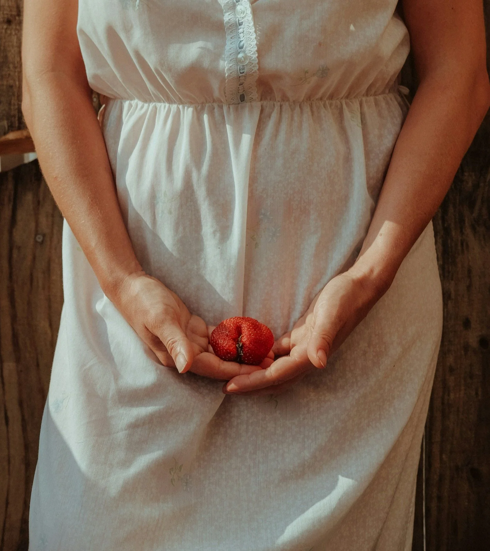 Une personne portant une robe blanche tient une fraise dans ses mains, assise devant un fond en bois.