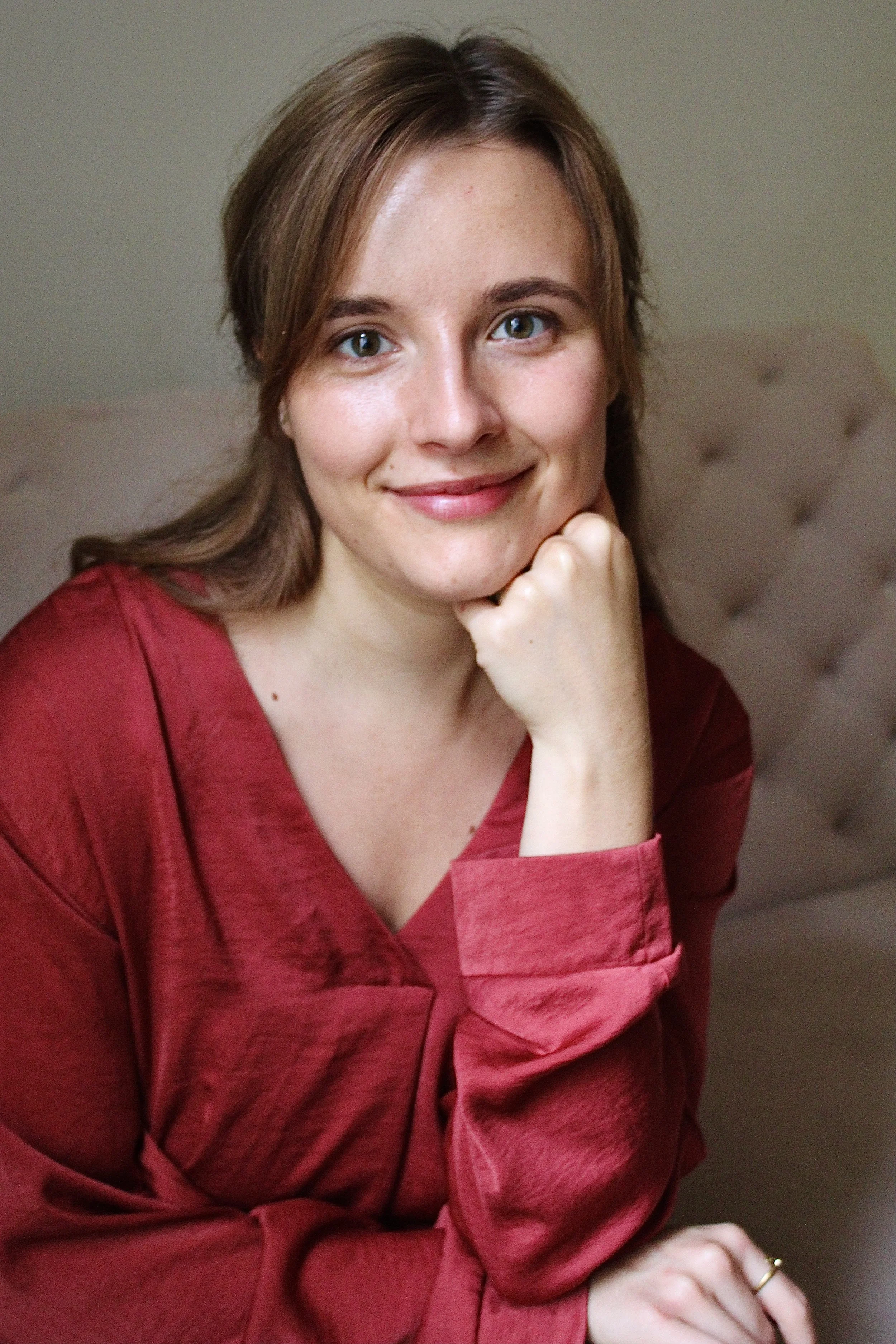 Photo professionnelle de Maude Guévin, femme bienveillante, souriante et inclusive avec une chemise rouge avec la main sous le menton, assise sur un canapé beige.