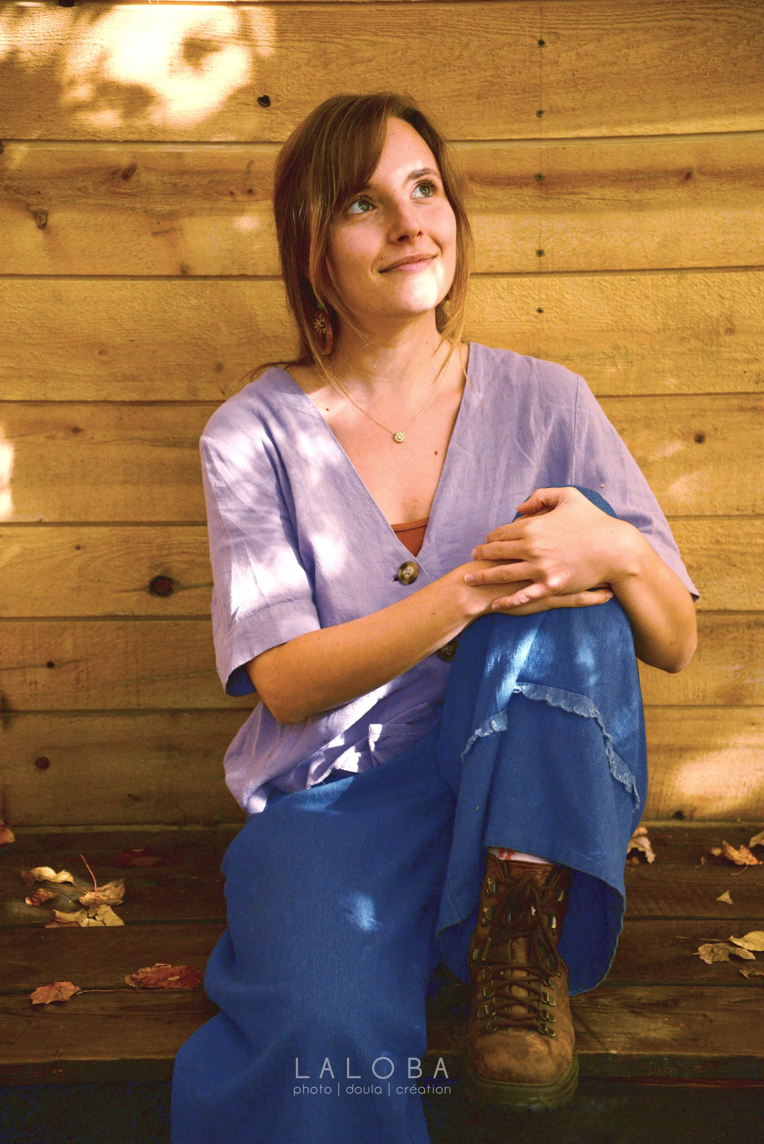 Une femme assise devant un mur en bois dans une pose pensive, portant une chemise violette et une jupe bleue, avec des bottes marron. Des feuilles mortes sont dispersées autour d'elle.