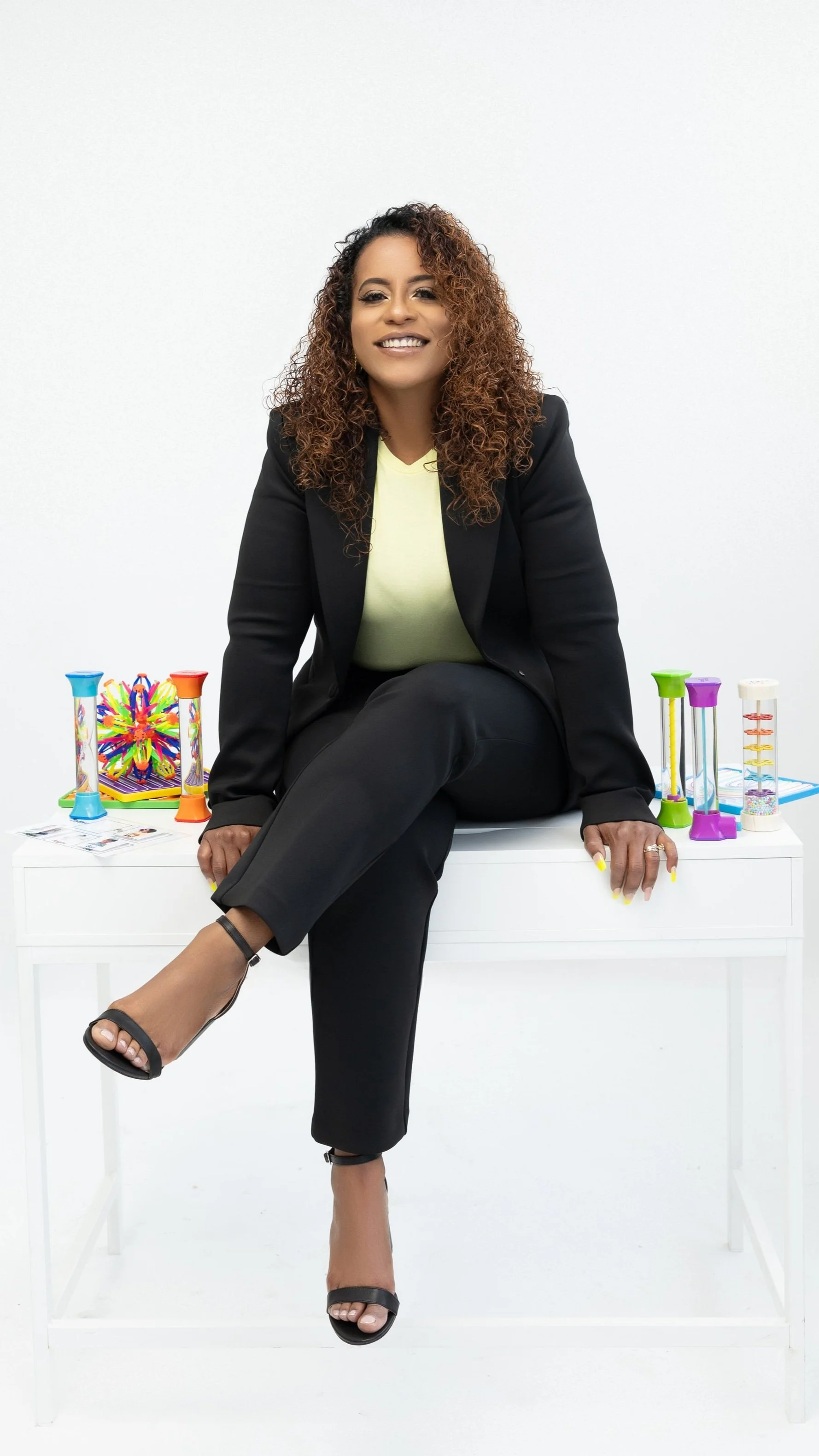 Mujer con cabello rizado y vestido amarillo sentado en una mesa blanca, rodeada de juguetes de diferentes colores.