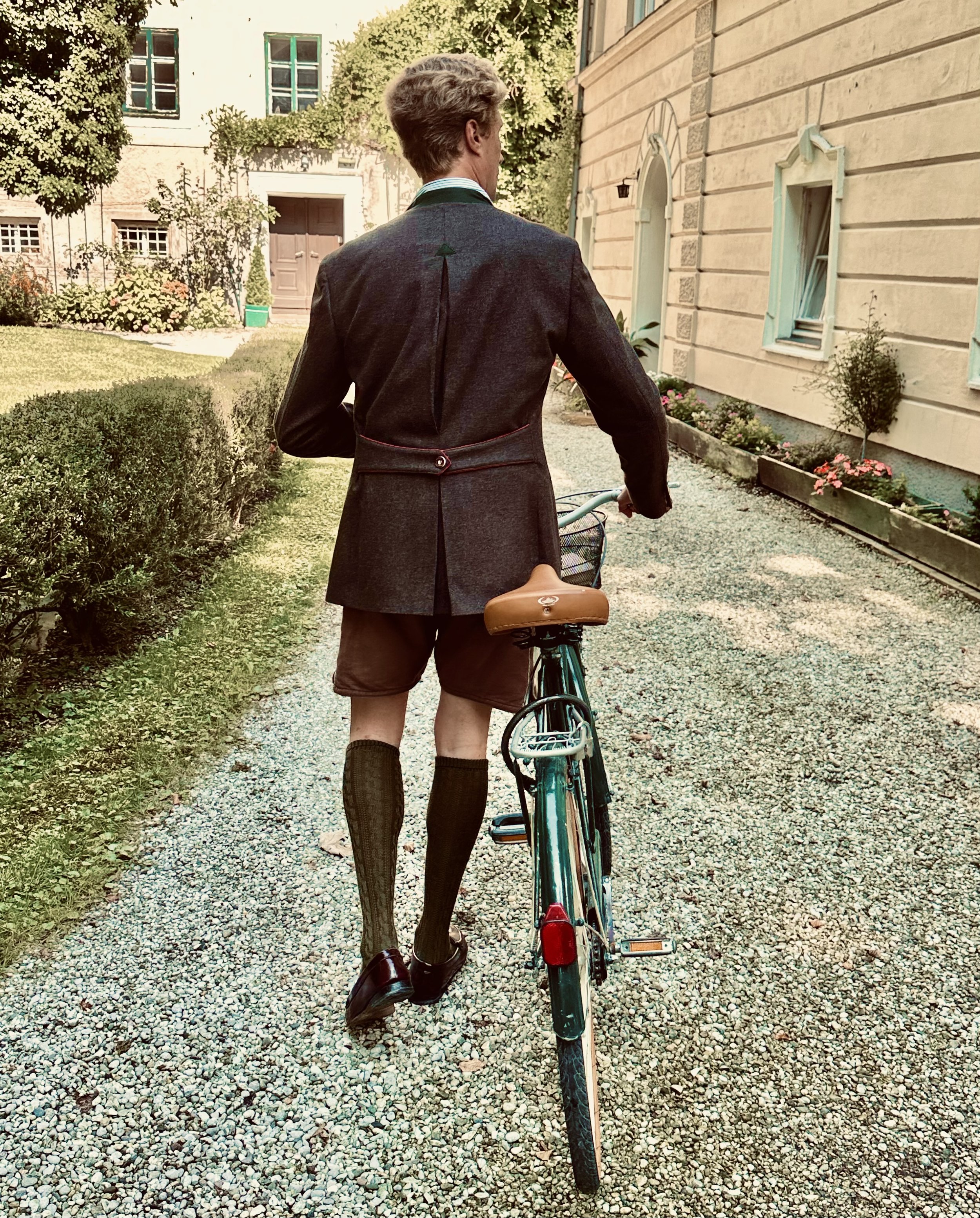 Giampiero Celani en lederhosen traditionel, portant de hautes chaussettes et des chaussures en cuir, pousse un vélo sur un chemin pavé devant une maison ancienne avec des fenêtres à volets. Le cadre est un jardin avec des plantes fleuries.