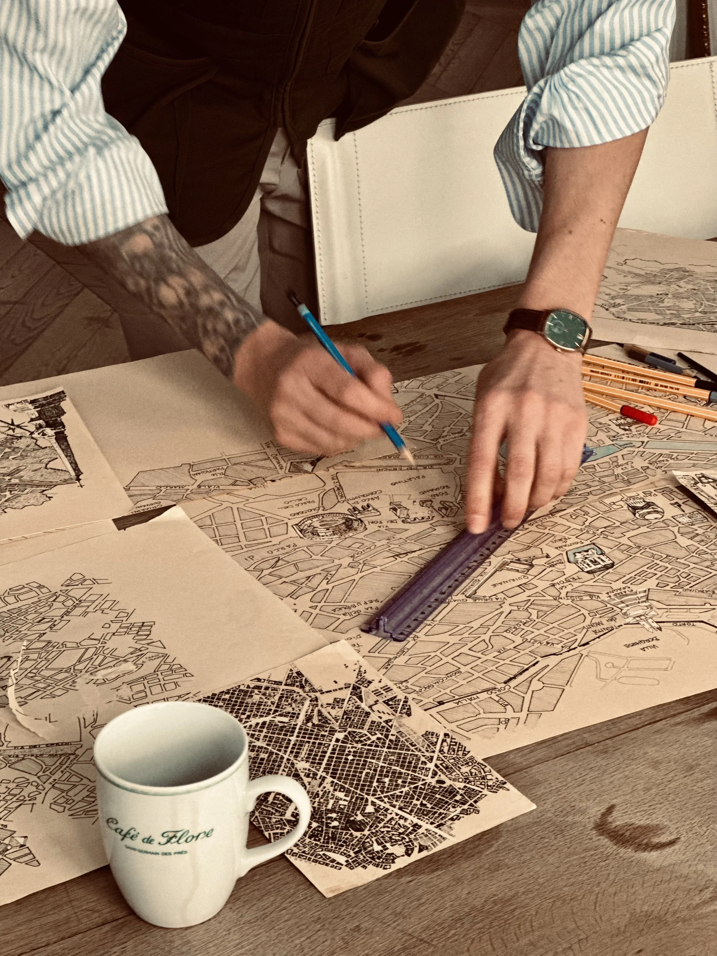 Personne tenant un stylo et une règle, en train de travailler sur une carte ancienne ou un plan dans une salle, avec une tasse et des crayons sur la table.