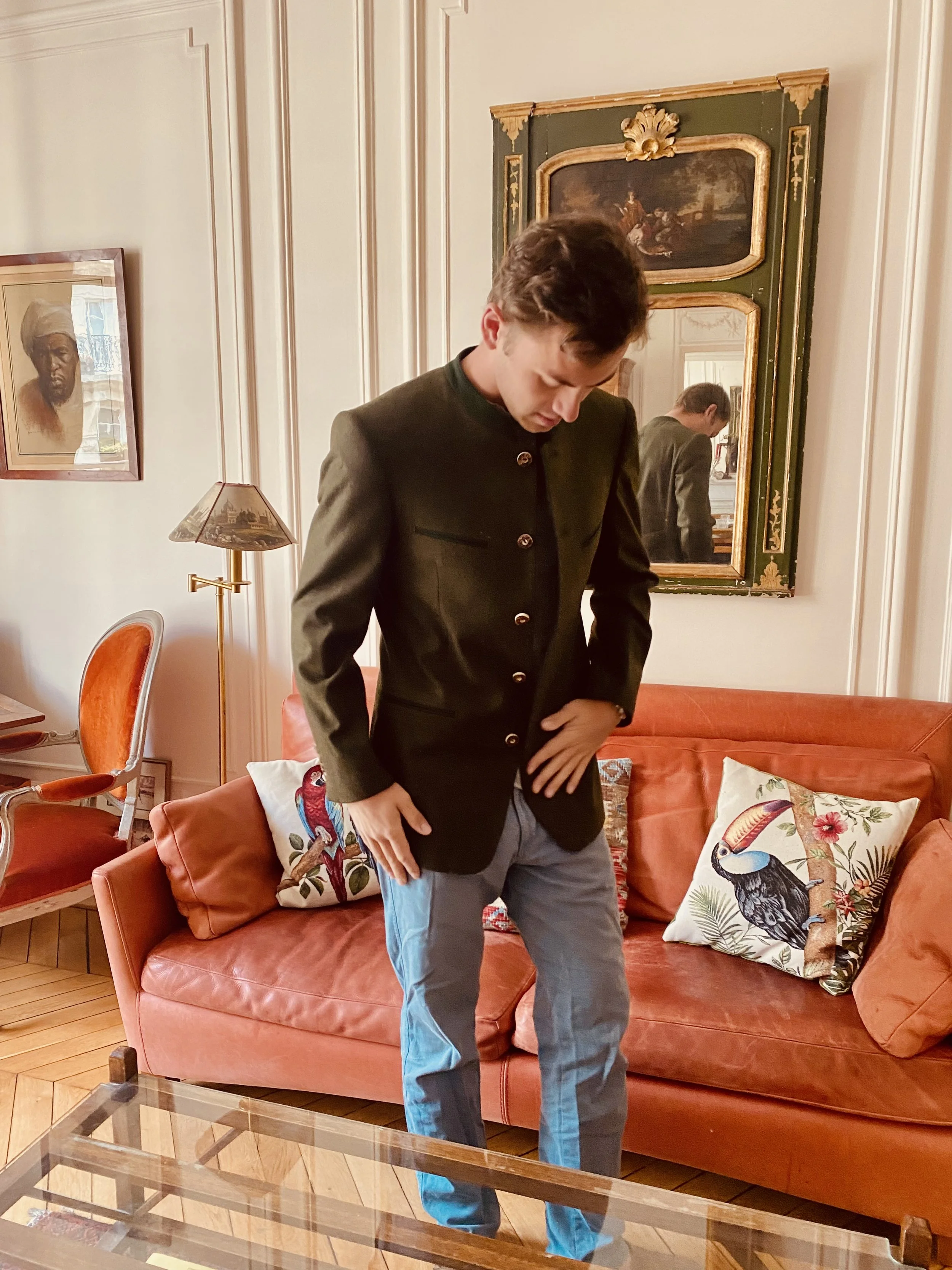 Jeune homme en veste sombre et pantalon gris dans un salon avec canapé orange, coussins à motifs de perroquets, murs ornés de tableaux et miroir.