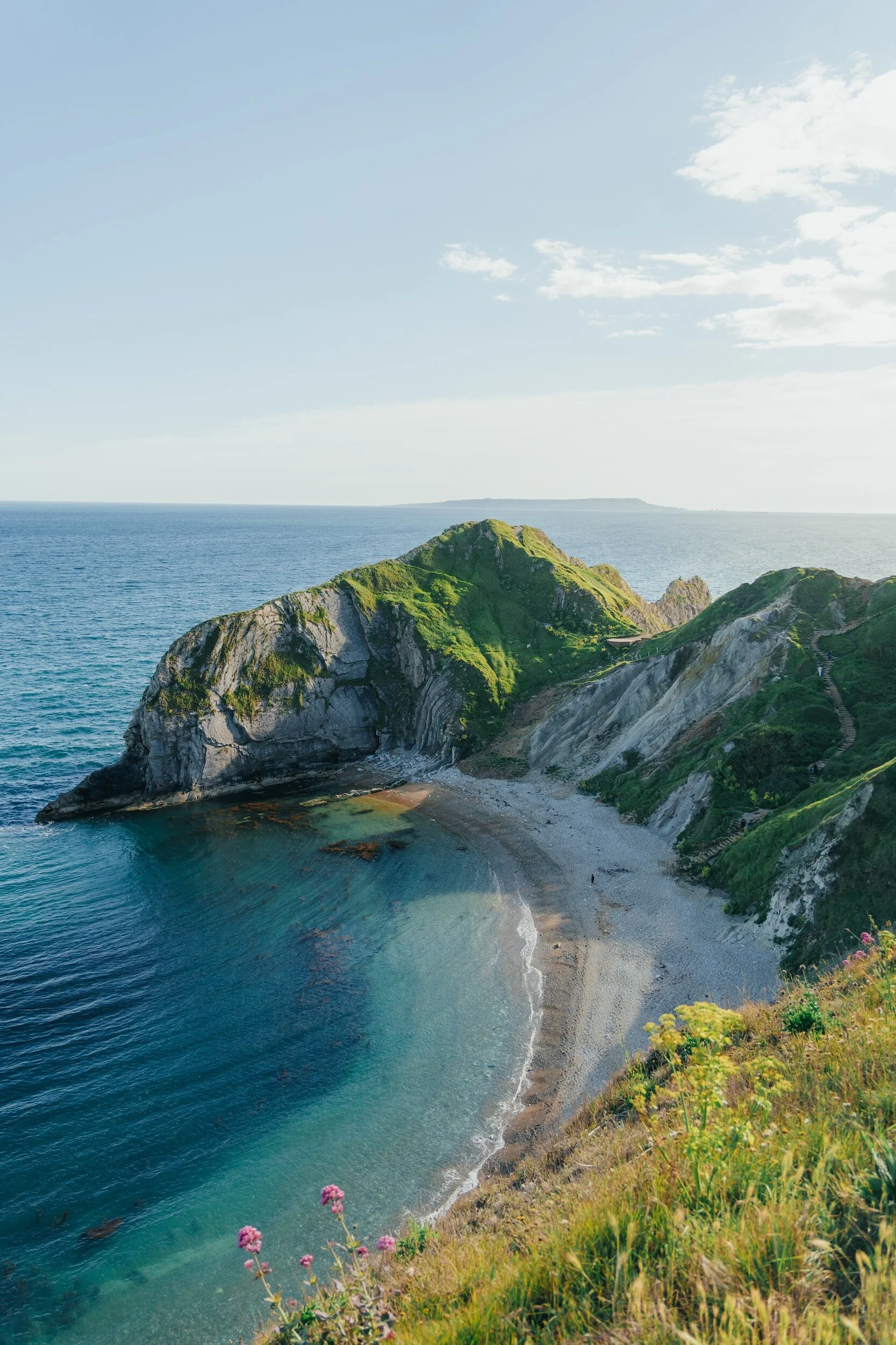 Scenic view of a green hilly coastal area with a small beach, clear blue water, and a partly cloudy sky.