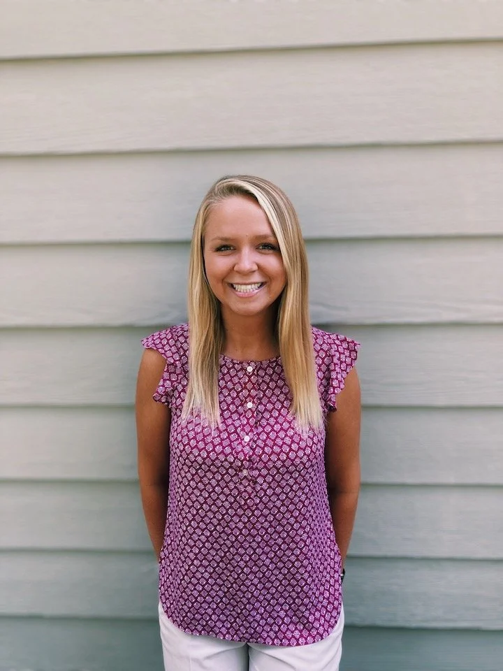 Smiling young woman with long blonde hair standing outdoors in front of a light gray horizontal siding wall, wearing a purple patterned sleeveless top and white pants.