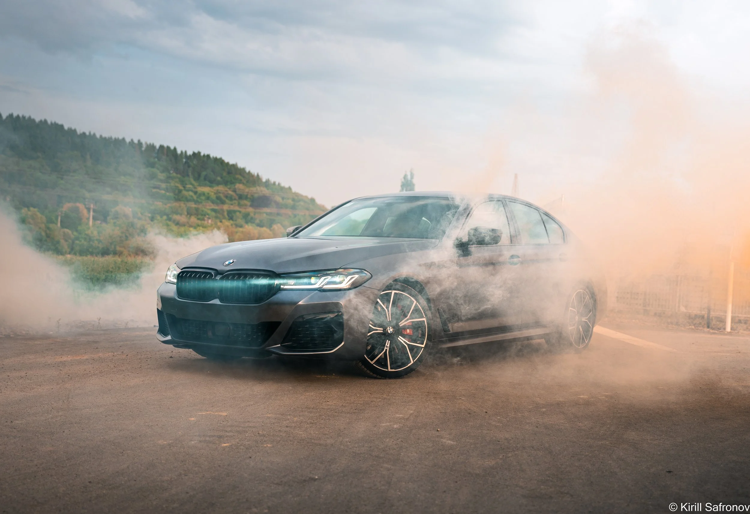 Photo of a grey BMW M550i driving on a rural road, kicking up dust, with a cloudy sky and countryside landscape in the background.