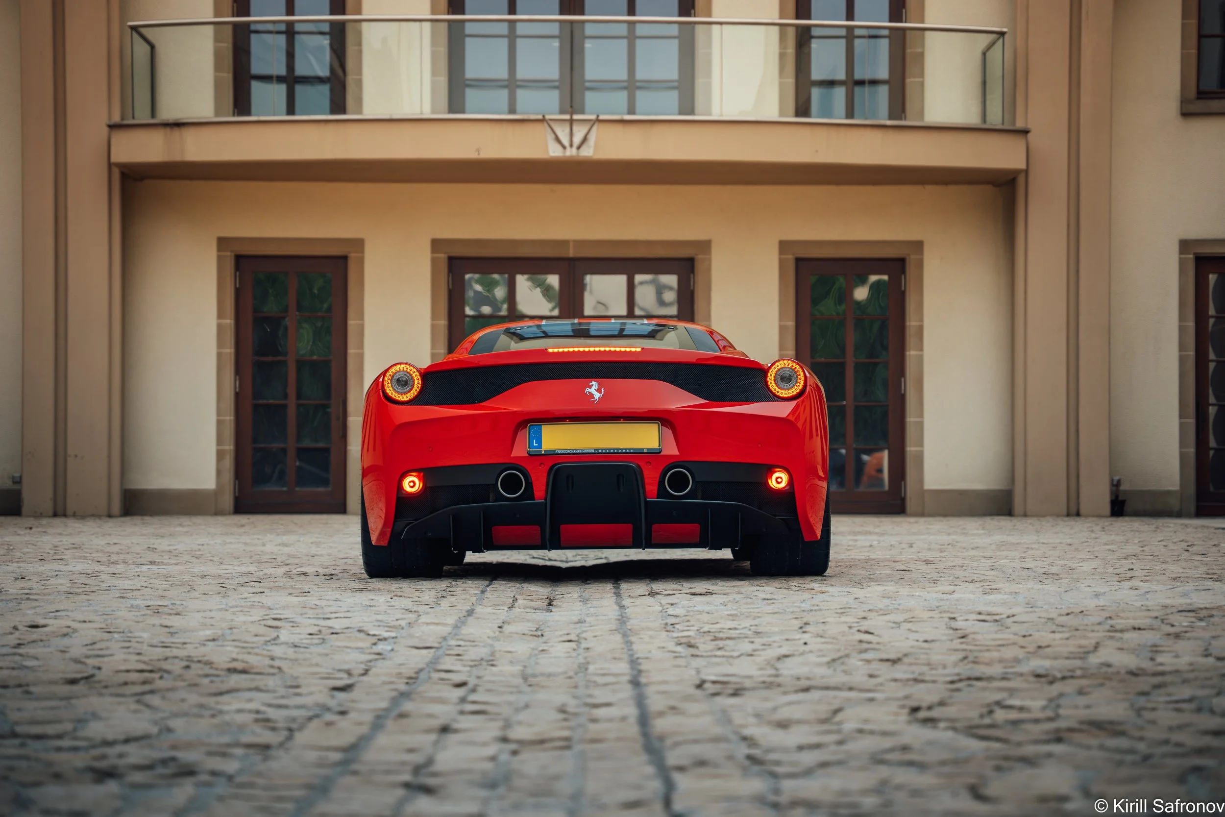 Rear view of a red Ferrari 458 Speciale parked outside a modern luxury house with a balcony and large glass doors.