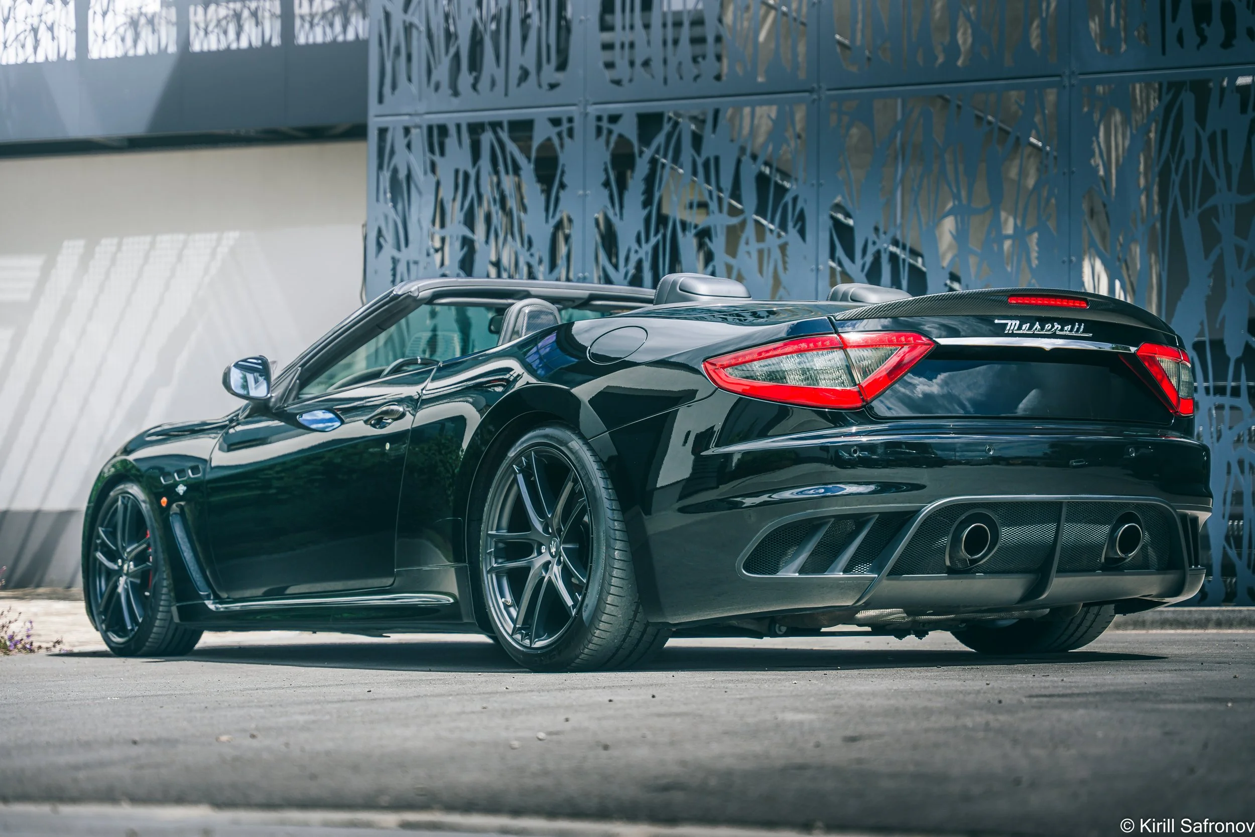 Photo of a black Maserati GranTurismo convertible parked in front of a modern metal building with abstract architectural patterns.