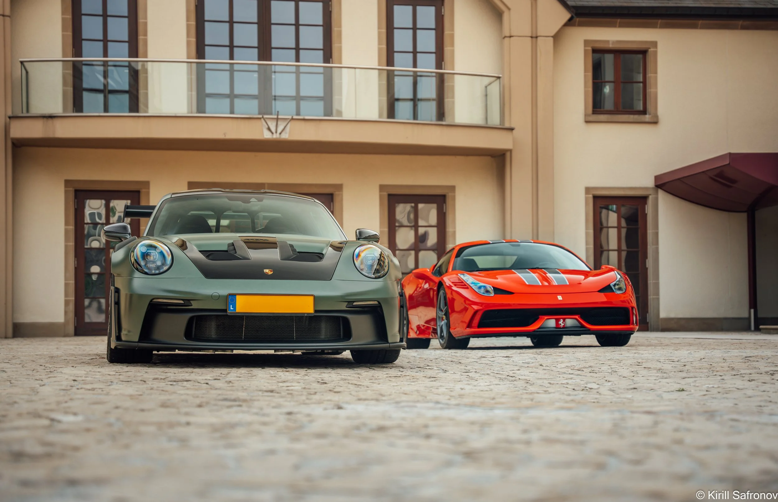 A dark green Porsche 911 GT3 RS and a red Ferrari 458 Speciale parked side by side outside a modern residential building with glass windows and a balcony, showcasing two iconic luxury sports cars in an urban setting.