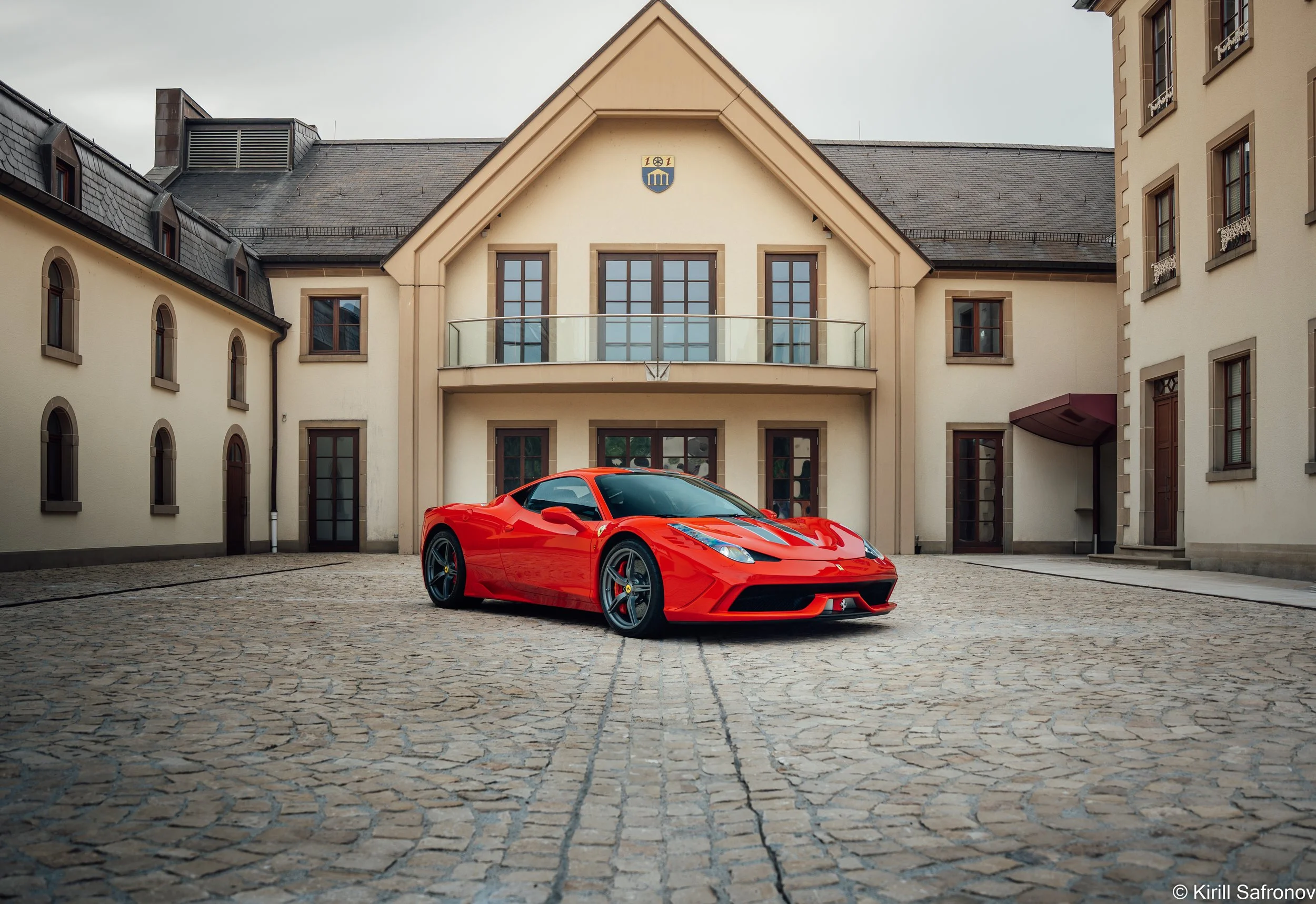 A red Ferrari 458 Speciale sports car parked on a cobblestone square in front of a residential building with classic windows and balconies, captured in an urban street photography style.