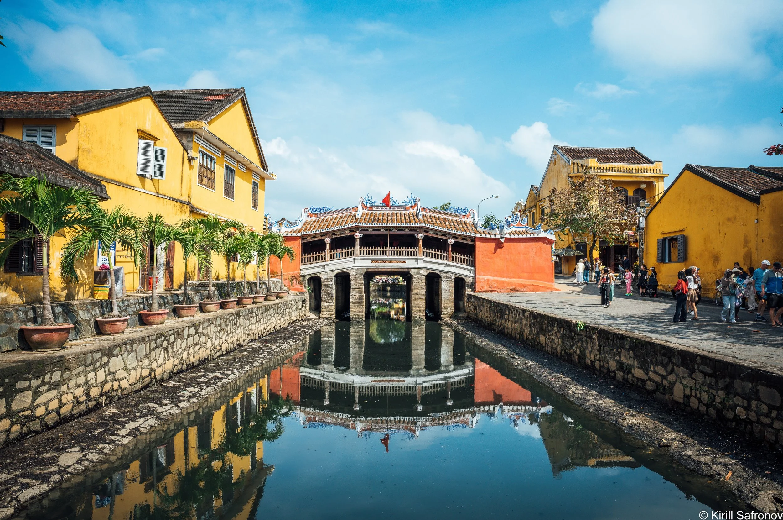 Photo of Chùa Cầu, the Japanese Covered Bridge in Hoi An, Vietnam, reflected in a canal beside old yellow buildings, palm trees, and tourists exploring the historic town.