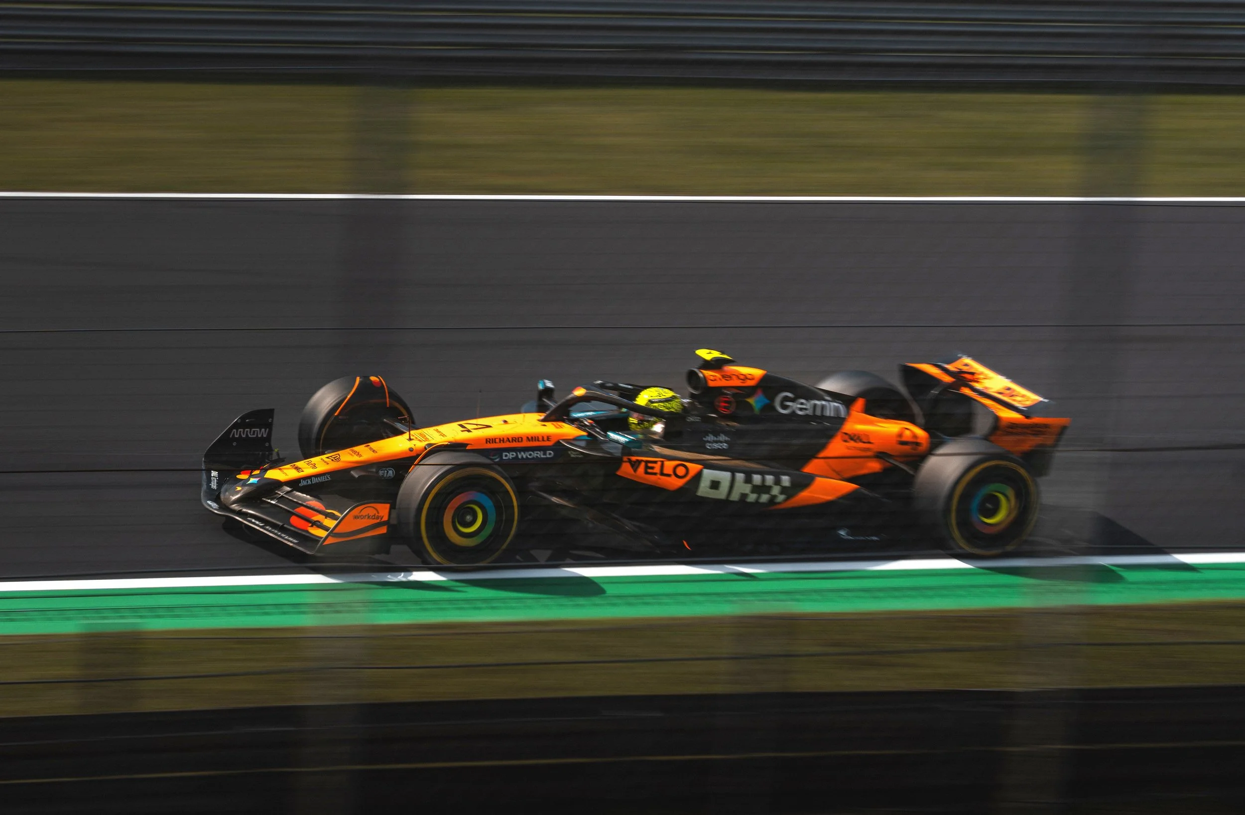 Photo of a McLaren orange Formula 1 car racing at high speed on the Monza circuit during the Italian Grand Prix, with dramatic motion blur and track safety barriers in the background.