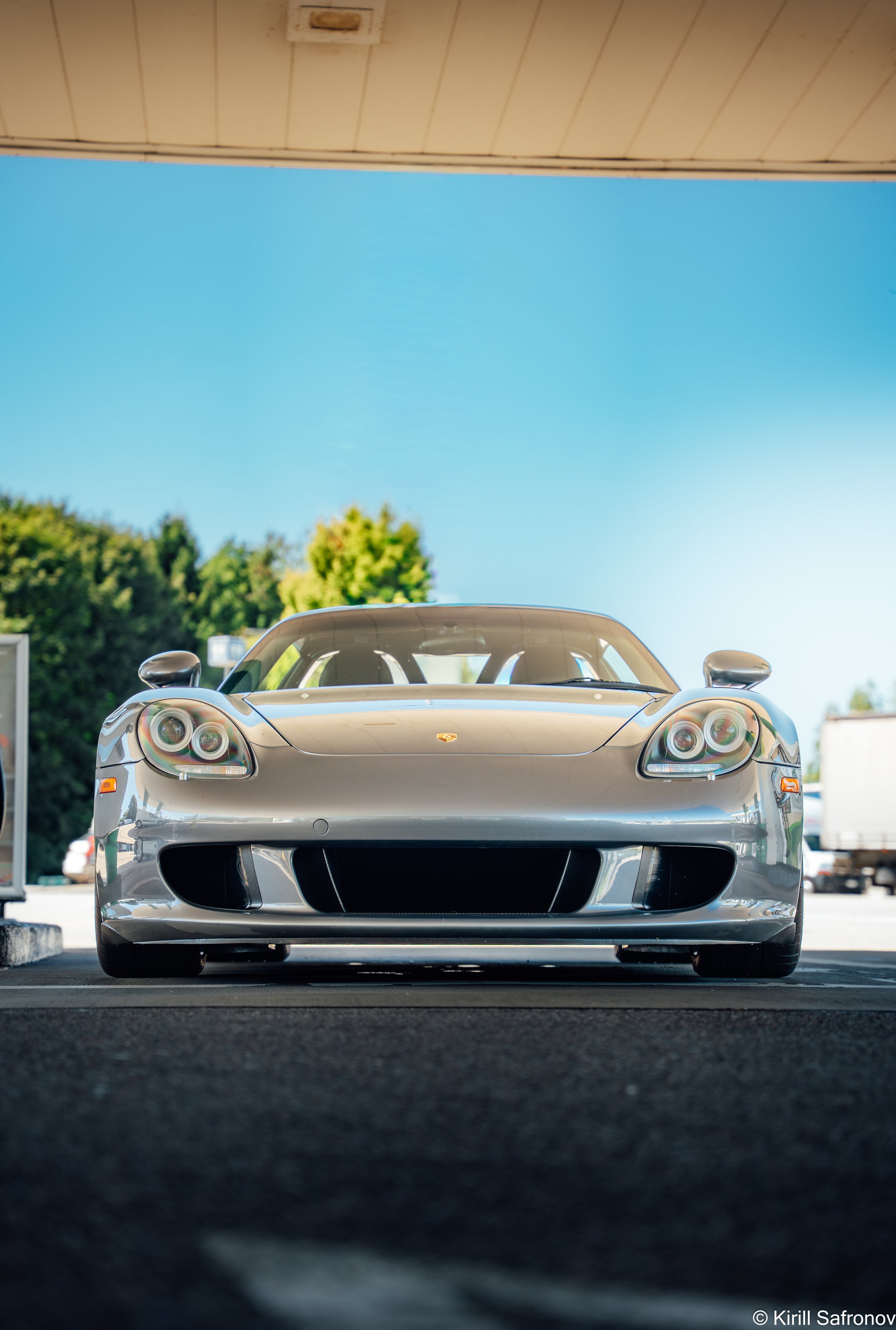 Front view photo of a silver Porsche Carrera GT sports car under a clear blue sky, with green trees in the background.