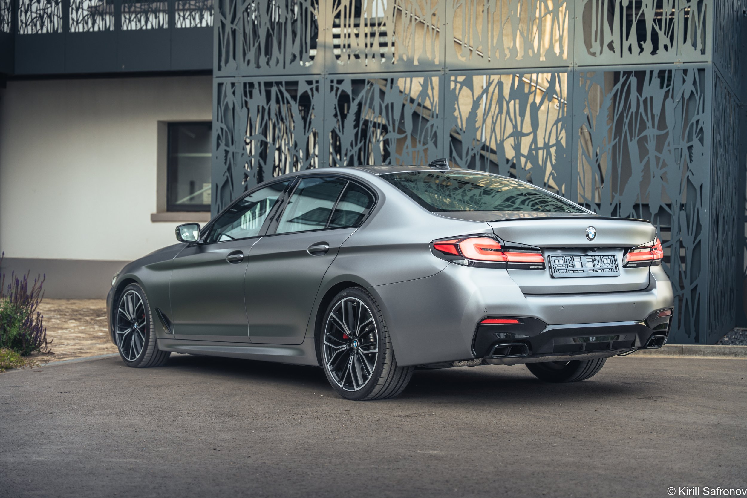 Photo of a grey BMW M550i rear view luxury sedan parked outside a modern architectural building with a decorative metal facade.