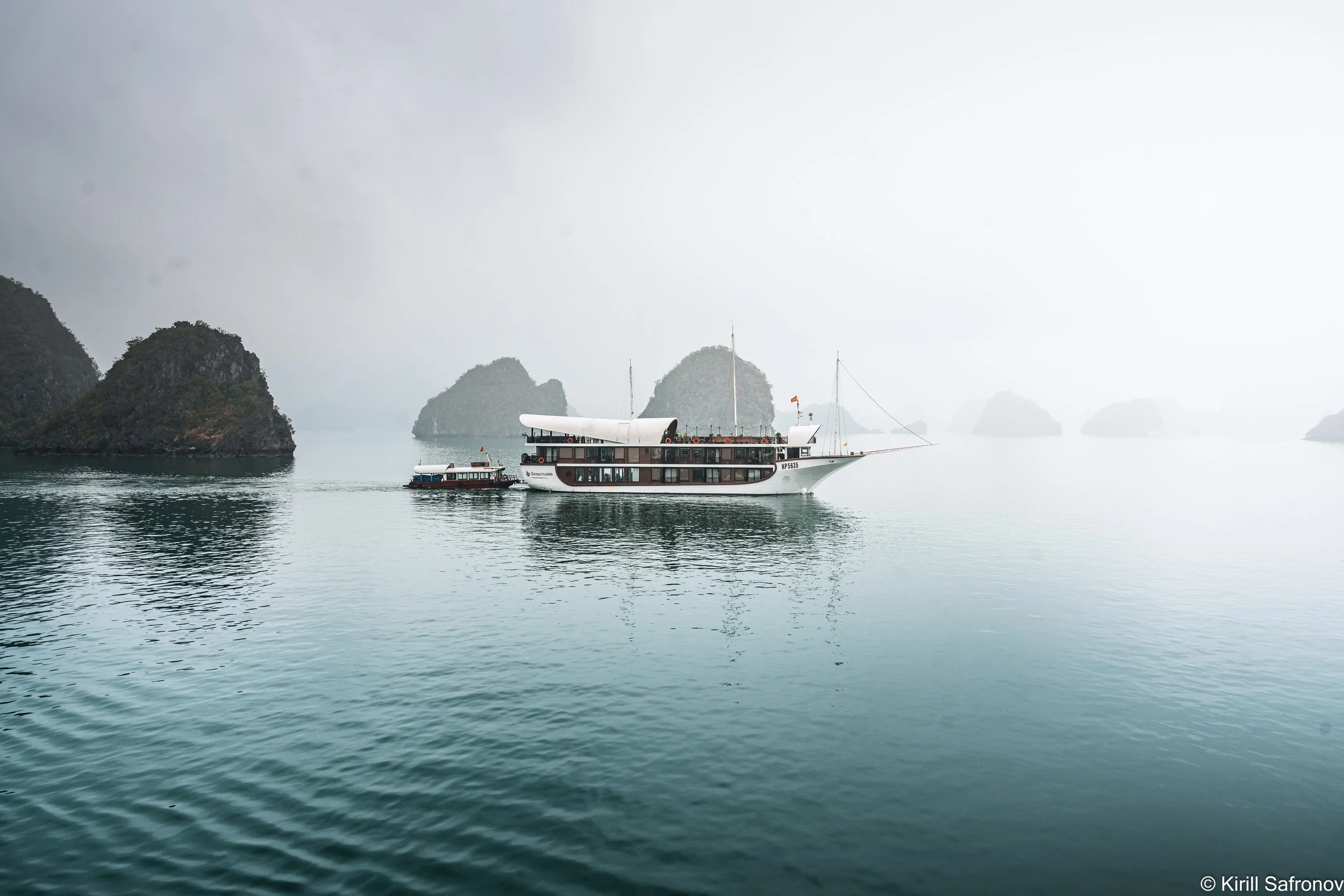 Photo of a traditional boat sailing across the calm waters of Ha Long Bay, Vietnam, surrounded by dramatic limestone karst islands, misty cliffs, and a hazy tropical sky.