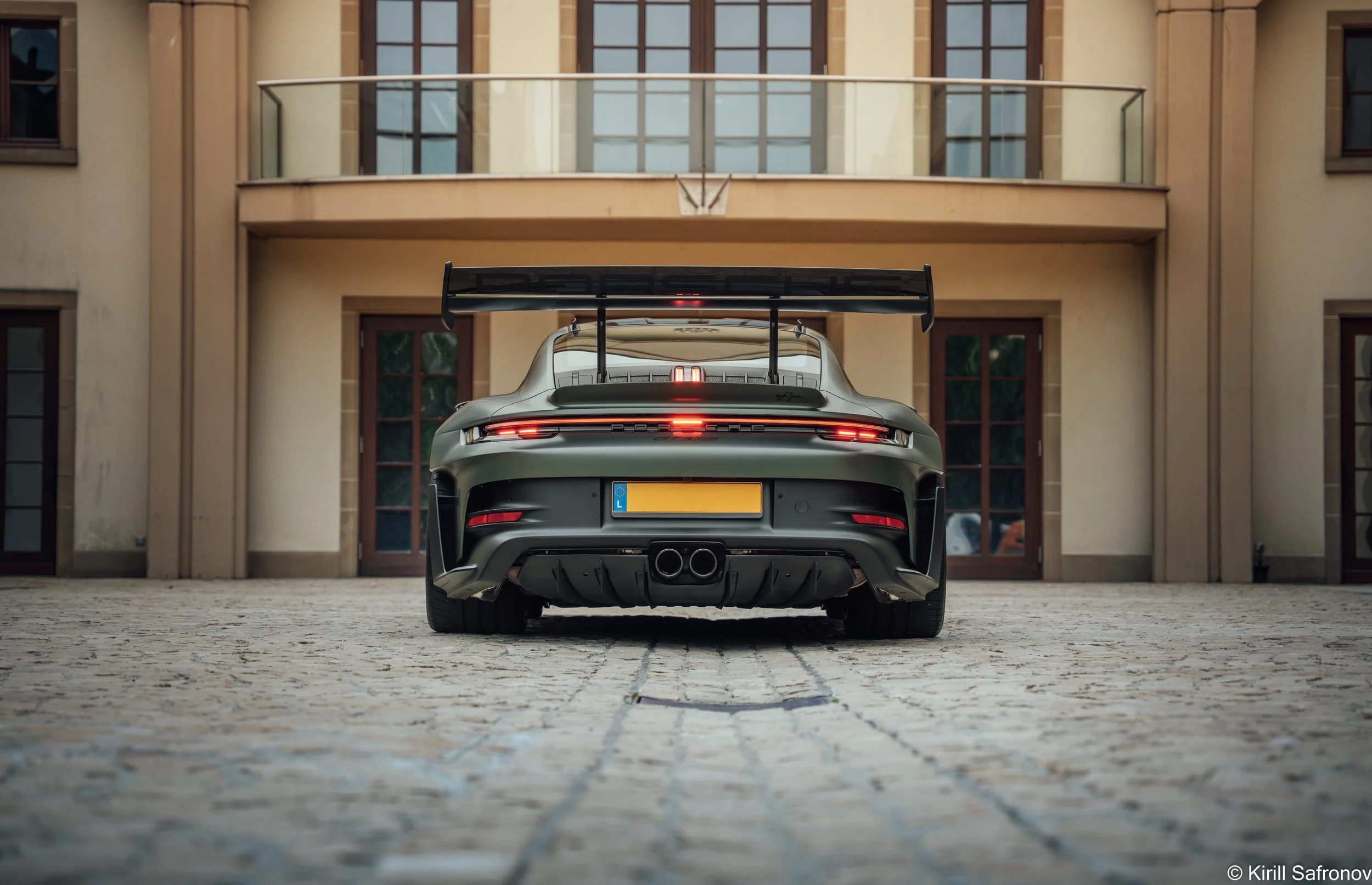 Rear view of a dark green Porsche 911 GT3 RS parked on a cobblestone driveway outside a modern luxury home with a glass gate.
