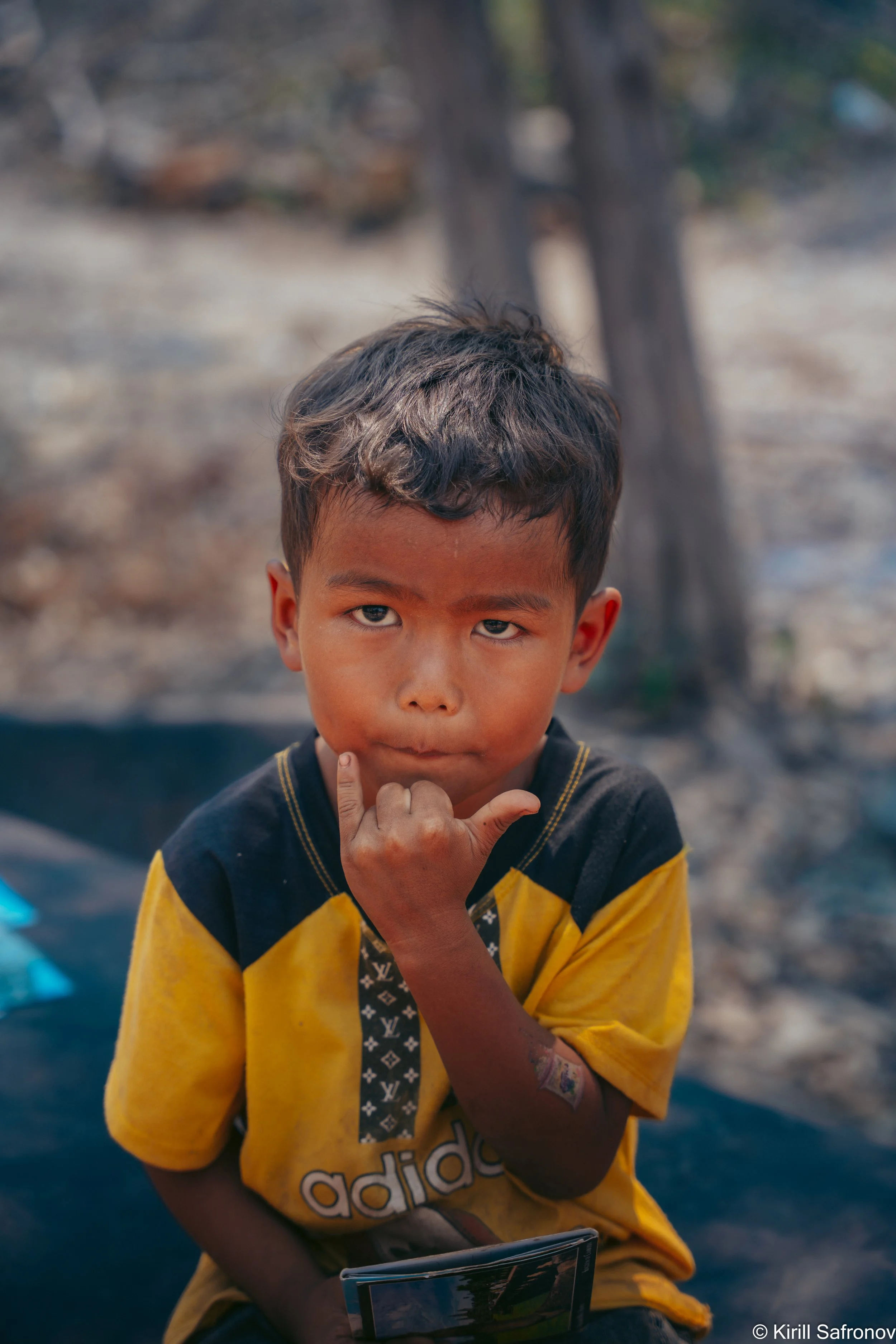 photo of a young boy sitting outdoors, wearing a yellow Adidas t-shirt, holding a tablet and looking at the camera, with a tree in the background.