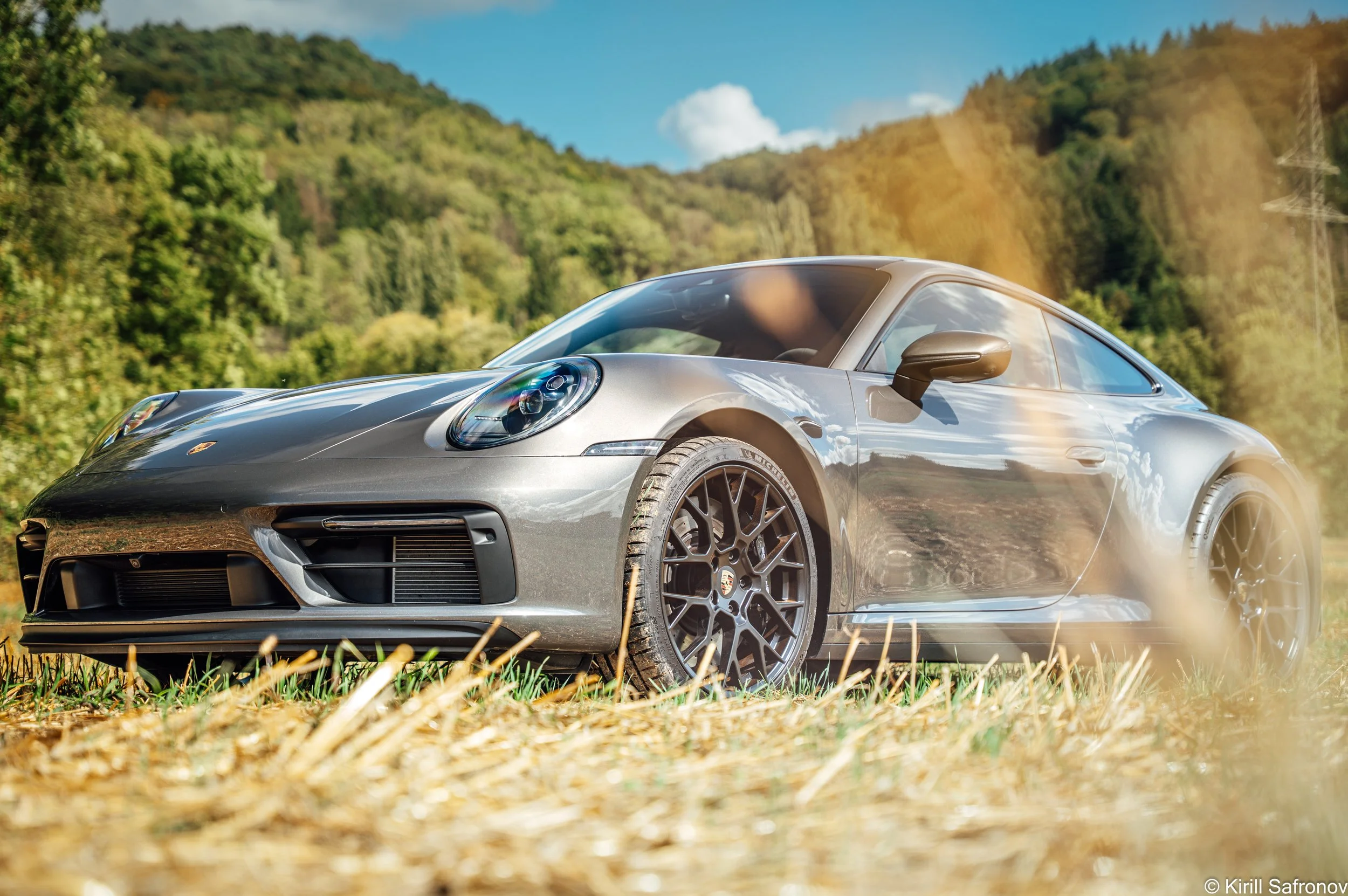 Champagne Porsche 911 Carrera GTS driving along a scenic countryside road, captured in motion against green hills and a bright blue sky.