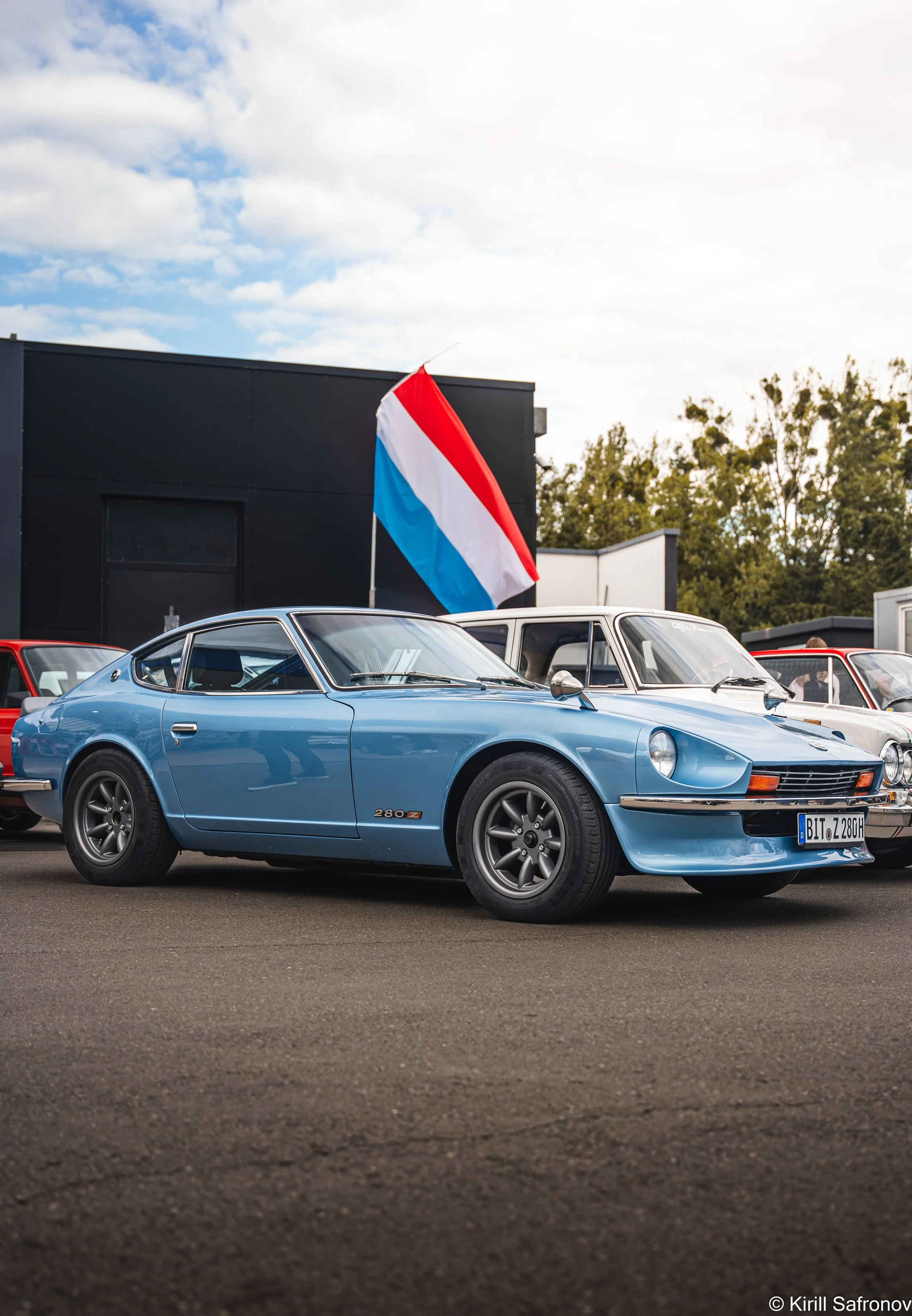 Photo of a blue 1976 Datsun 280Z parked outdoors with the Luxembourg flag in the background under a partly cloudy sky.