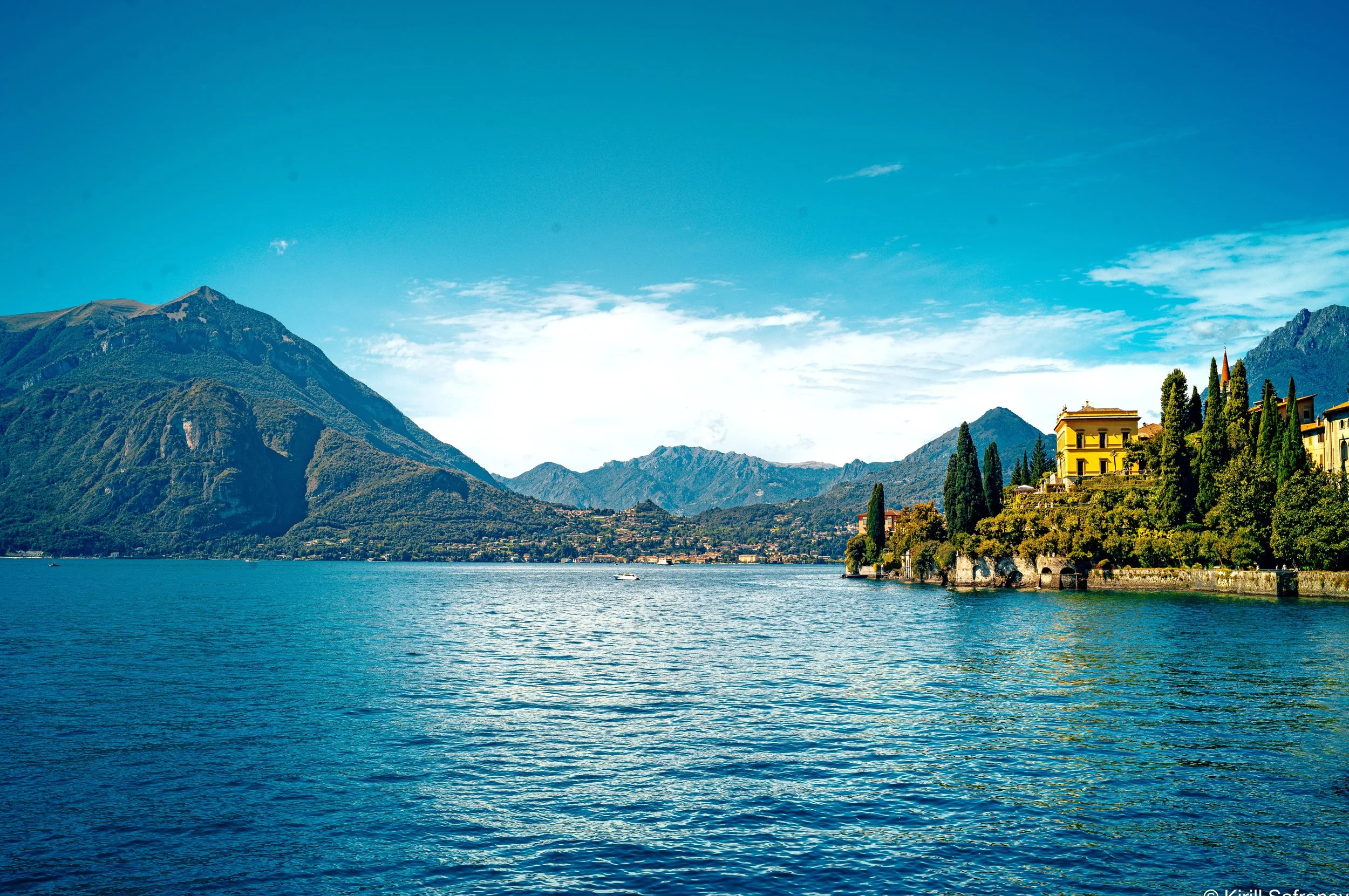 photo of Lake Como with mountains, lakeside buildings, calm blue water, and a bright sky with soft clouds in northern Italy
