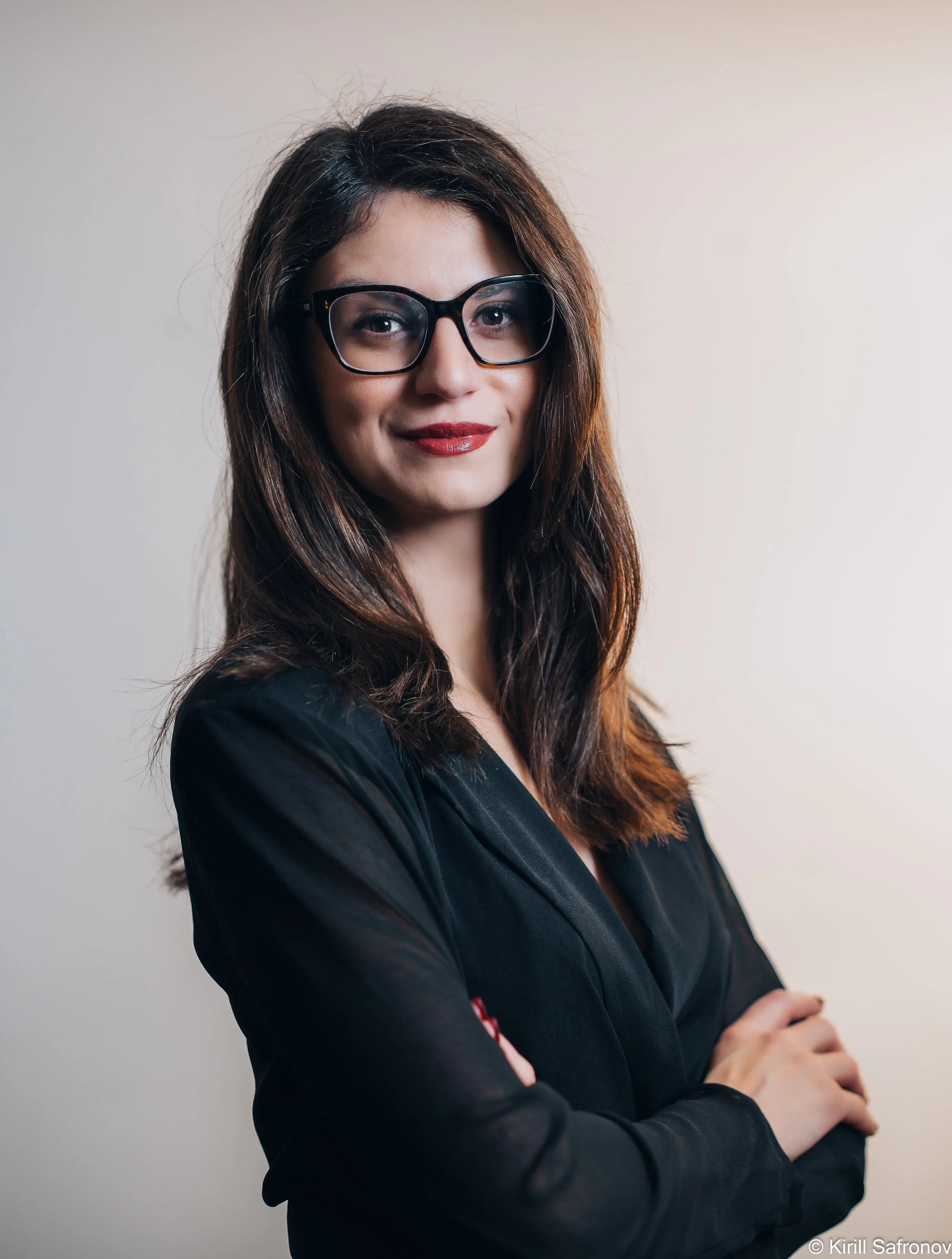 Photo of a smiling brunette woman with long hair wearing black glasses and a black jacket, posing with crossed arms against a light neutral background.