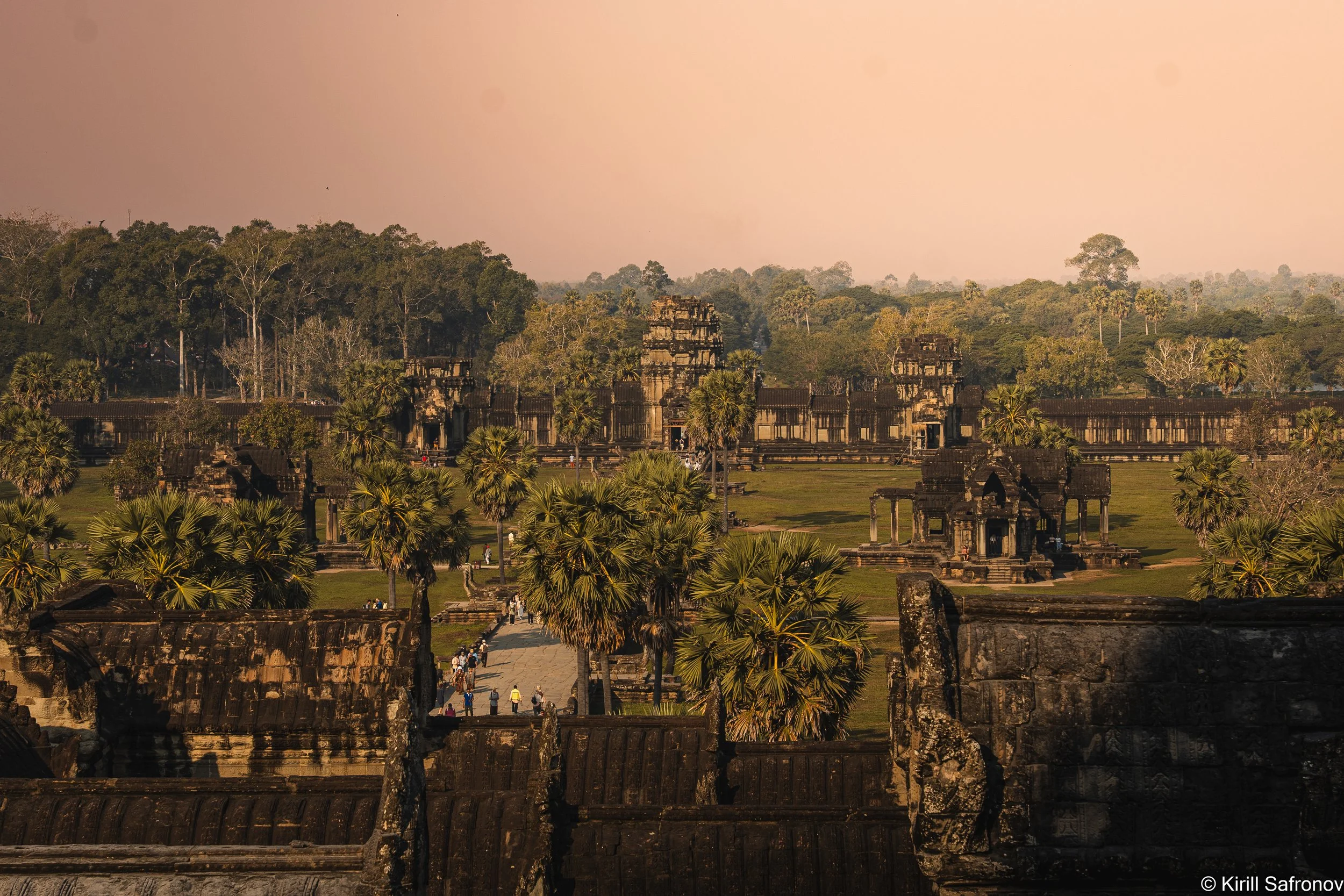 Photo of Angkor Wat temple enclosure in Cambodia, surrounded by dense jungle vegetation and old trees beneath a dark dramatic sky, creating a mysterious twilight atmosphere.