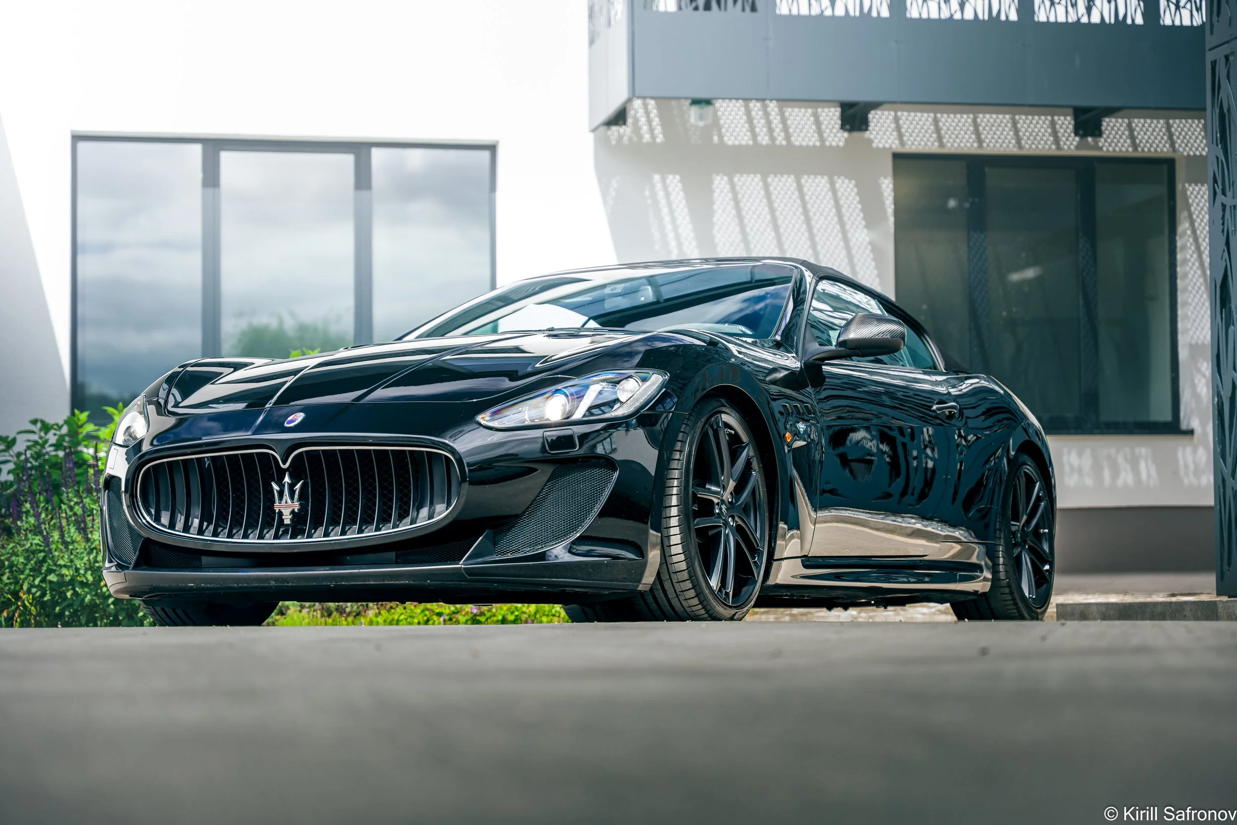 Photo of a black Maserati GranTurismo parked in front of a modern glass building under a cloudy sky.