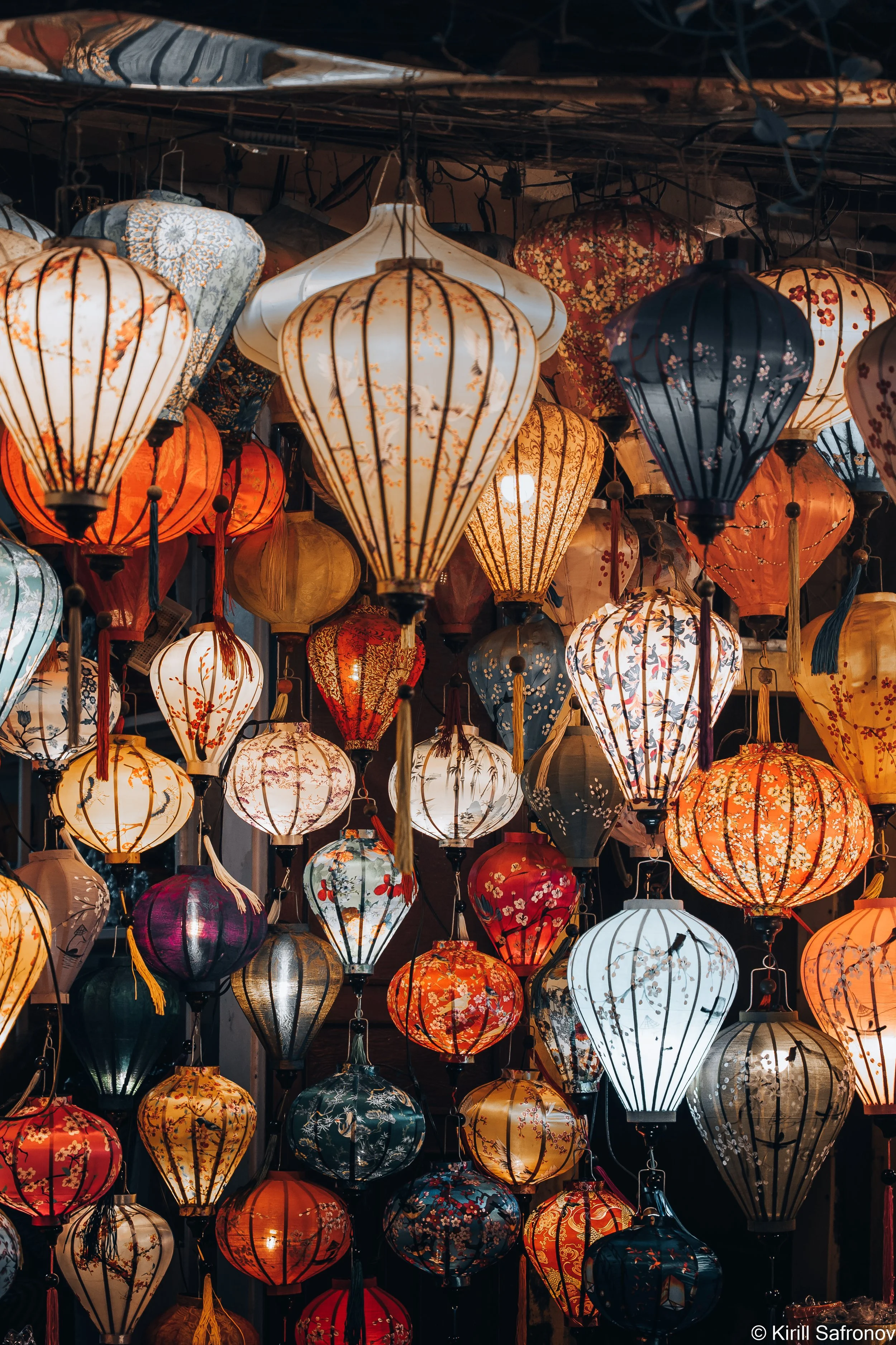 Photo of colorful hanging lanterns glowing at night, illuminating the scene with vibrant floral and geometric patterns in different shapes and sizes.