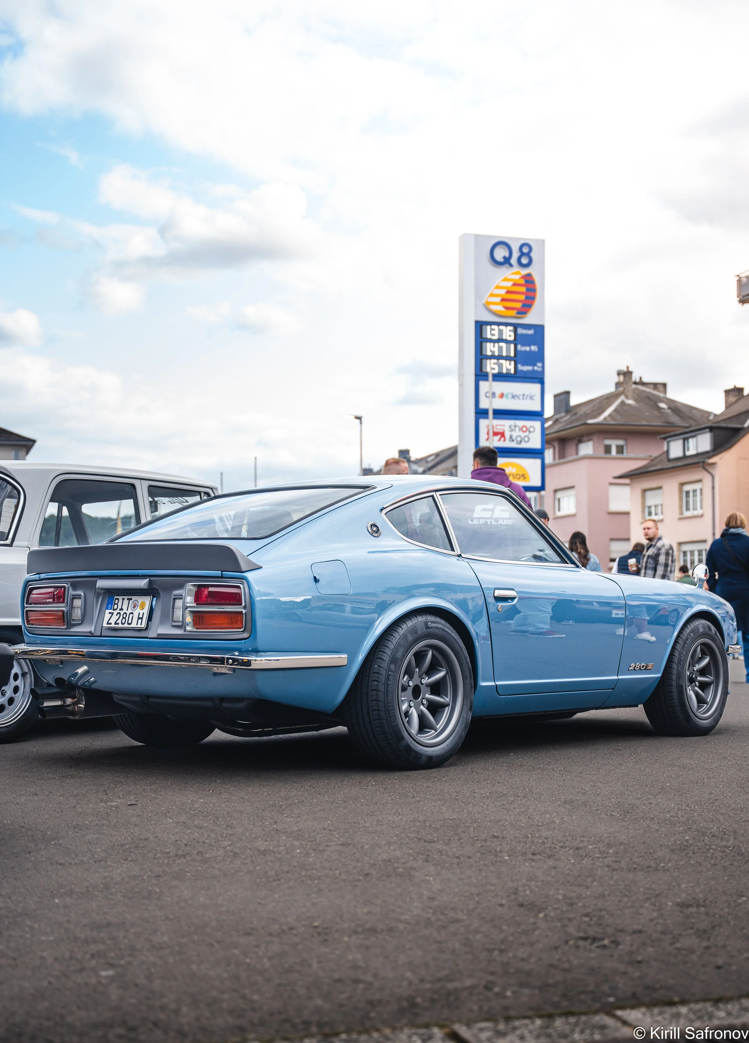 Photo of a blue 1976 Datsun 280Z parked on an urban street, with people in the background, residential buildings, a visible gas station sign, and a partly cloudy sky.