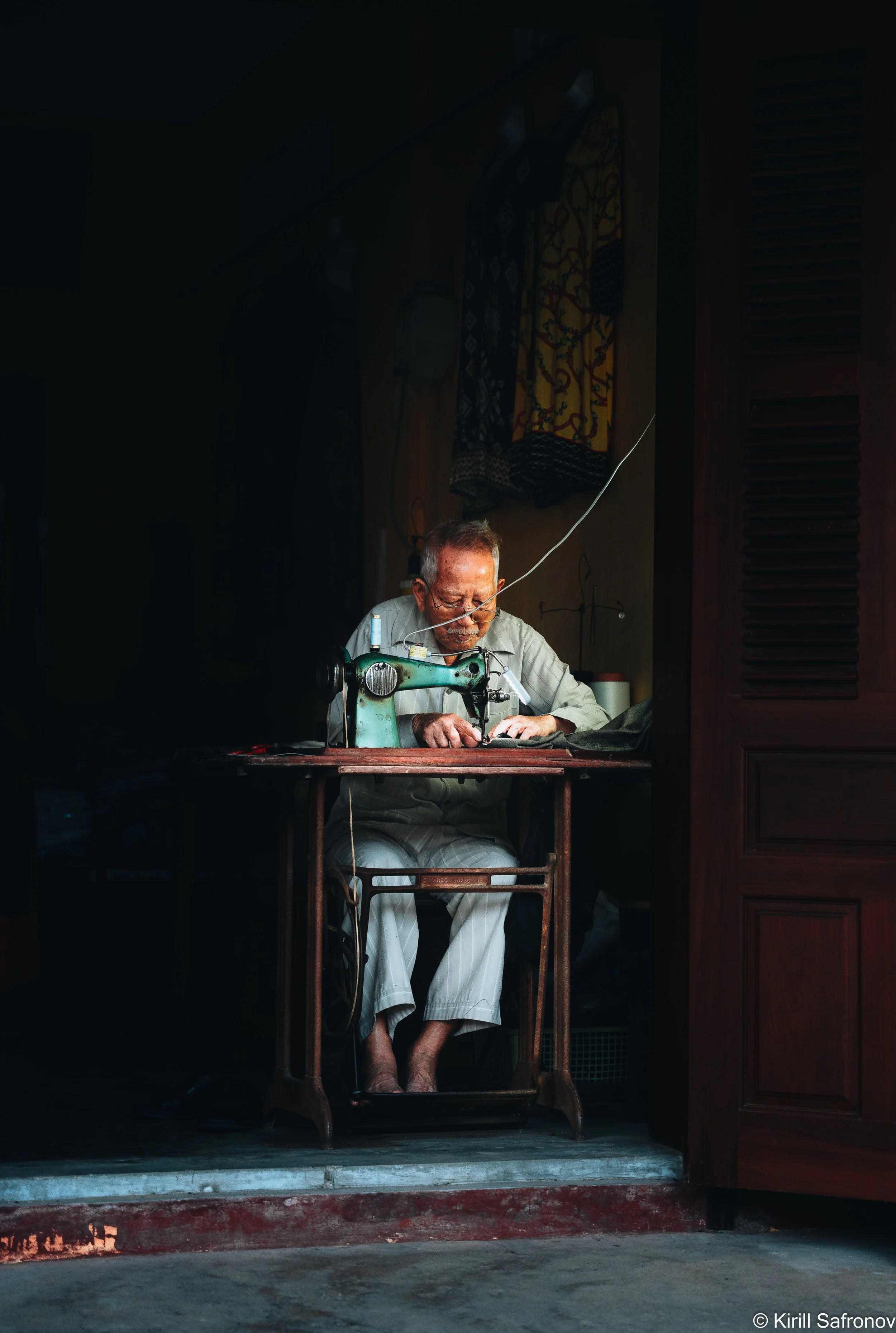 Photo of an elderly man with glasses sewing fabric on a vintage sewing machine in a dimly lit room, with dramatic focused lighting highlighting his craft and concentration.