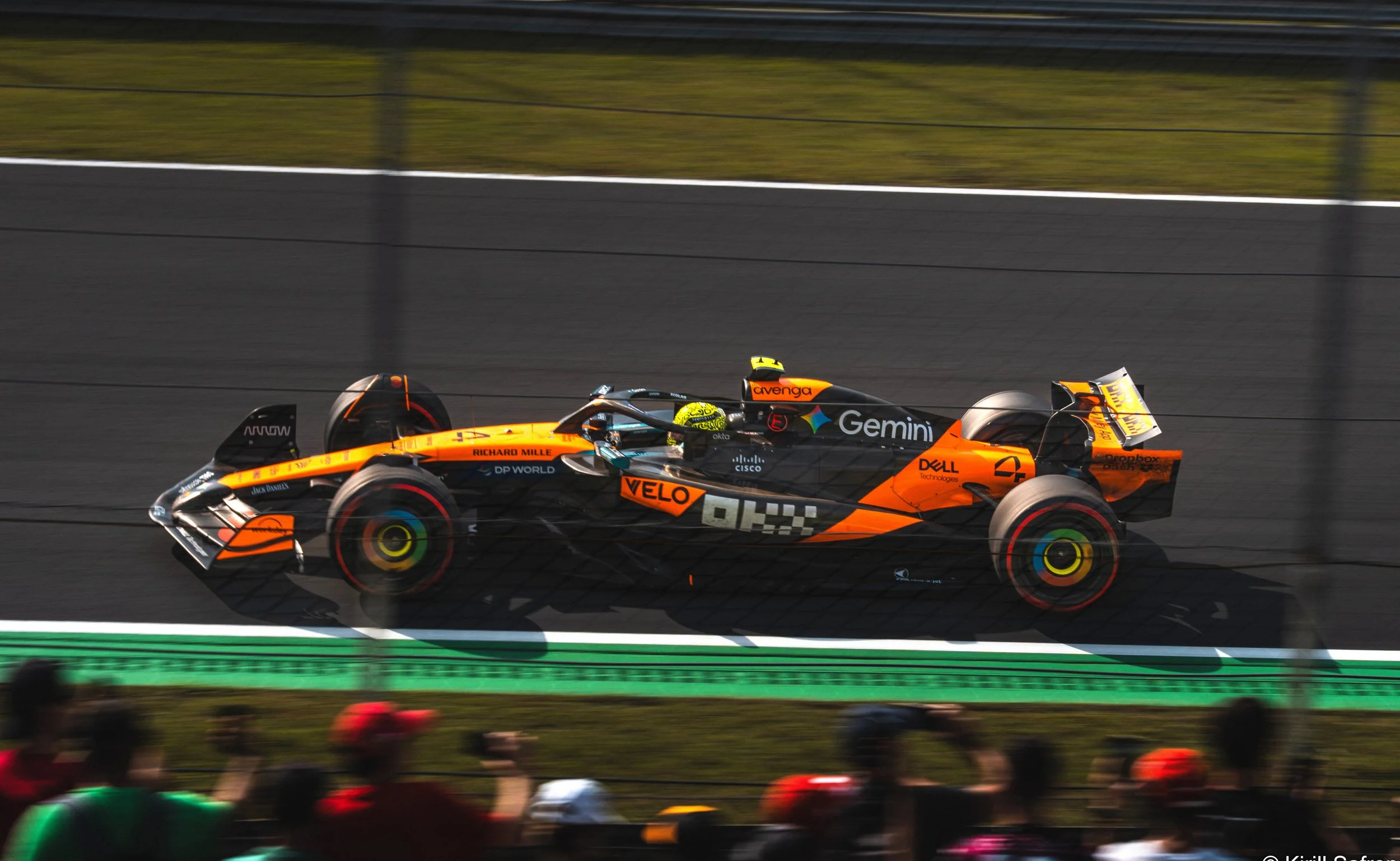 Photo of an orange McLaren Formula 1 race car speeding through the Monza Circuit during the Italian Grand Prix, with blurred spectators in the foreground capturing the energy and motion of the race.