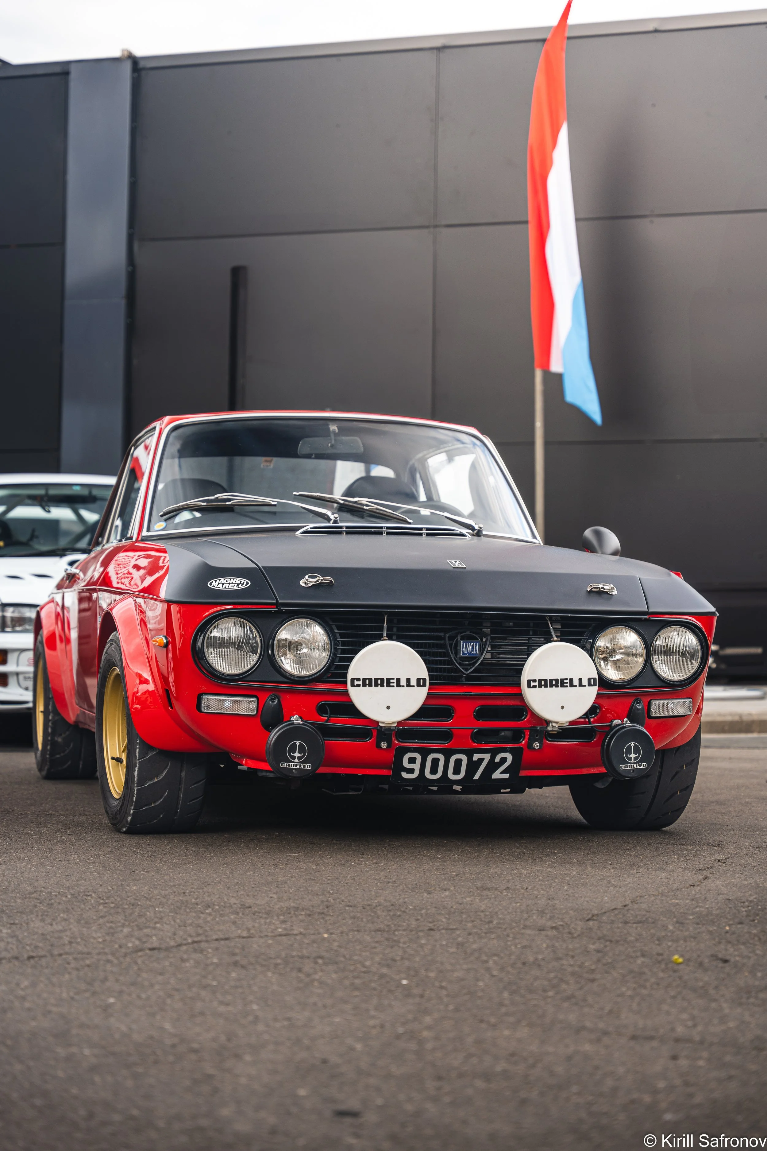 Photo of a classic red and black Lancia Fulvia 1.6 HF sports car with square dipped headlights, parked outside a grey building with a Luxembourg flag in the background.